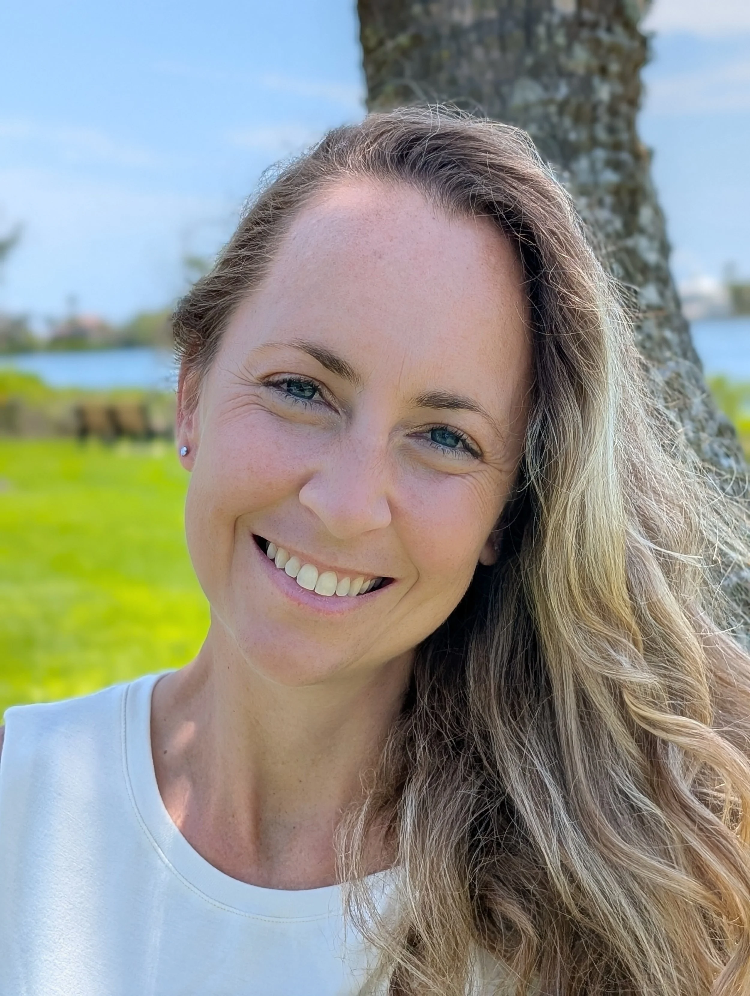 Close-up of a smiling woman with curly hair, blue eyes, and a white top outdoors near a tree and water.
