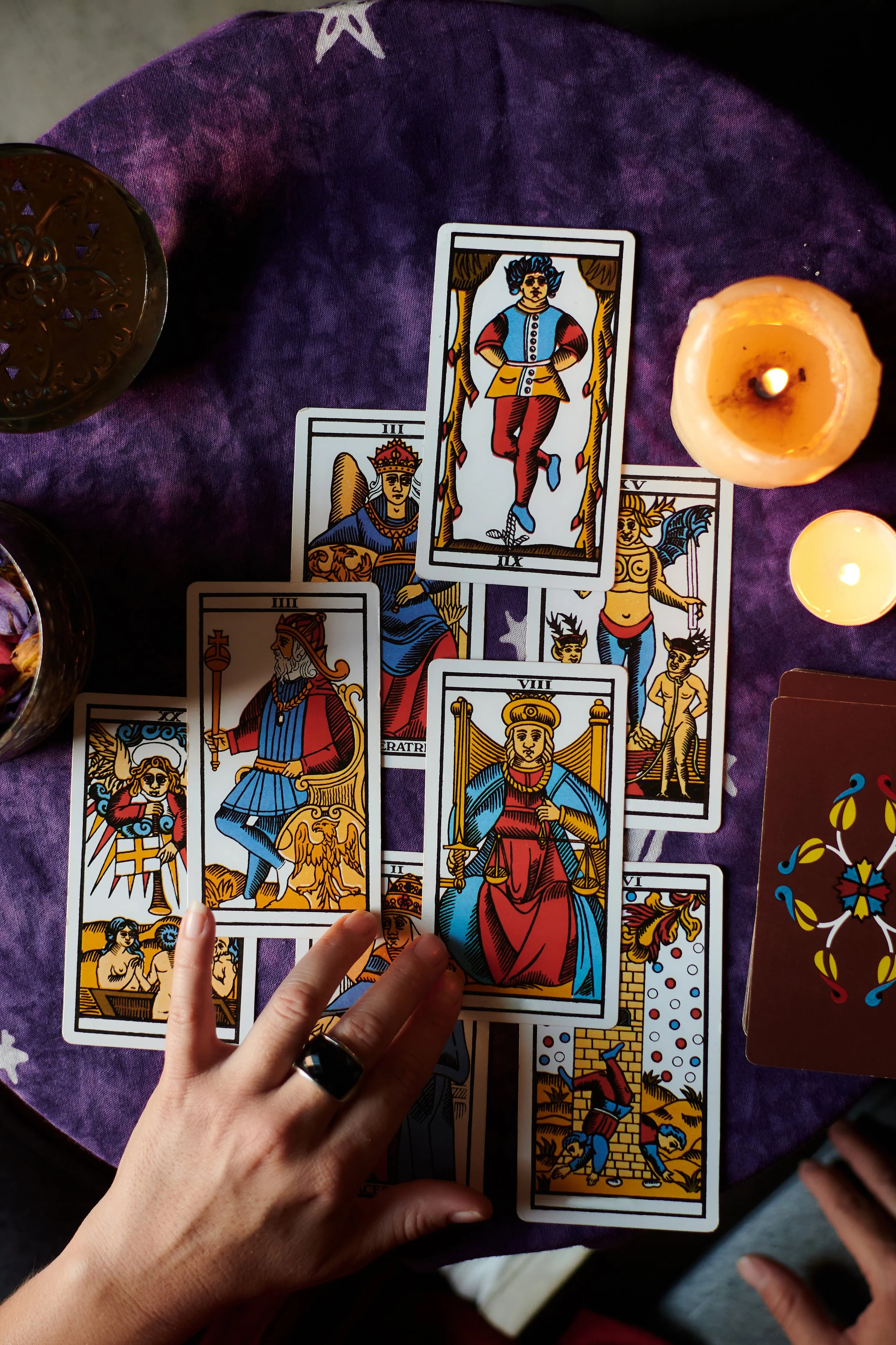 A spread of tarot cards on a purple cloth, surrounded by lit candles and a hand with a ring, with one card being held upright.