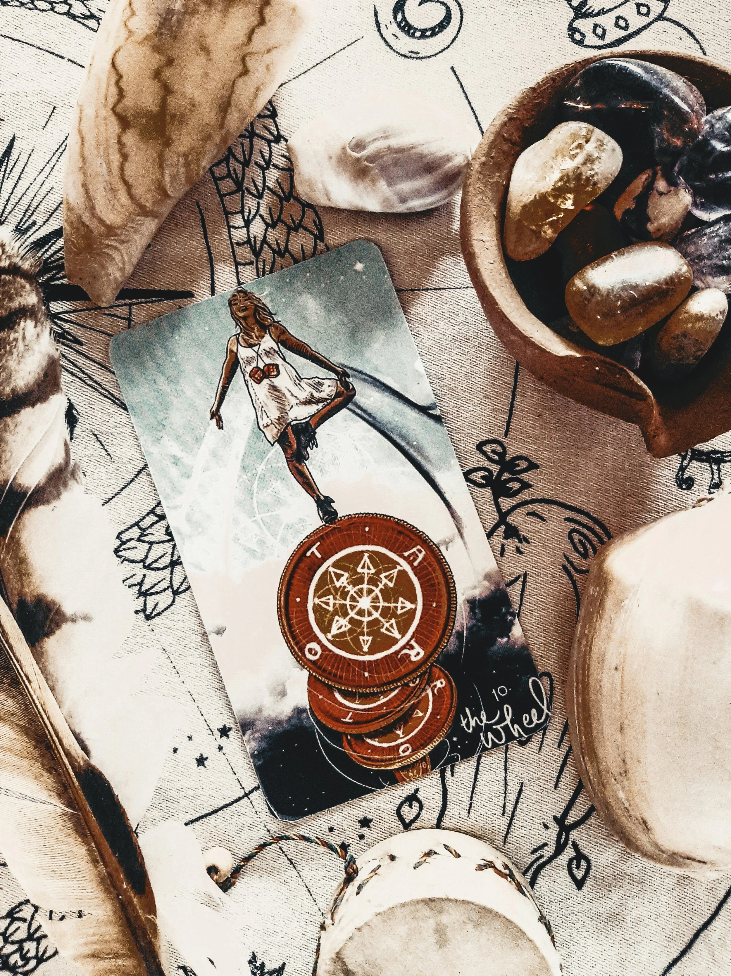 A tarot card with a woman in a white dress, surrounded by stones, shells, and decorative symbols on a patterned cloth, with a small container of gold-foil rocks.