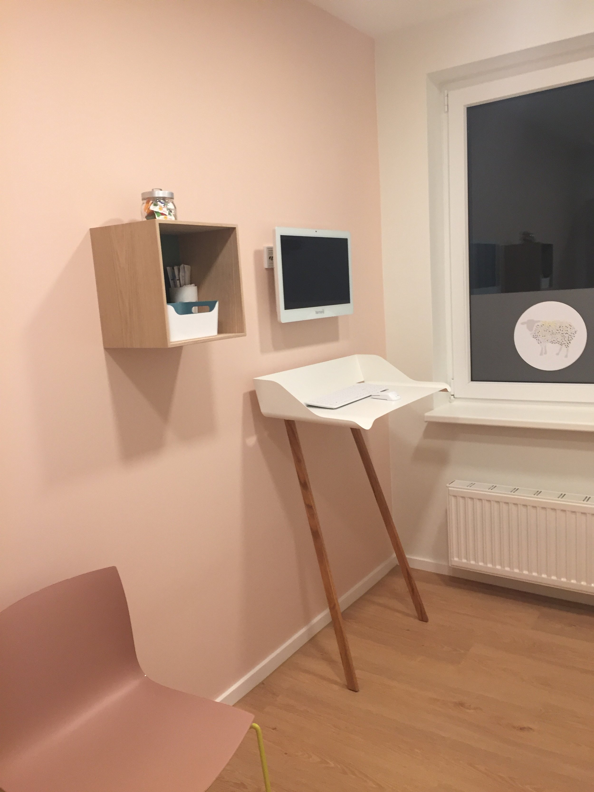 Ein Kinderzimmer mit rosa Wand, einem weißen Wickeltisch, einem kleinen schwarzen Bildschirm, einer hölzernen Wandschrank und einem Fenster mit einem Sheep-Aufkleber.
