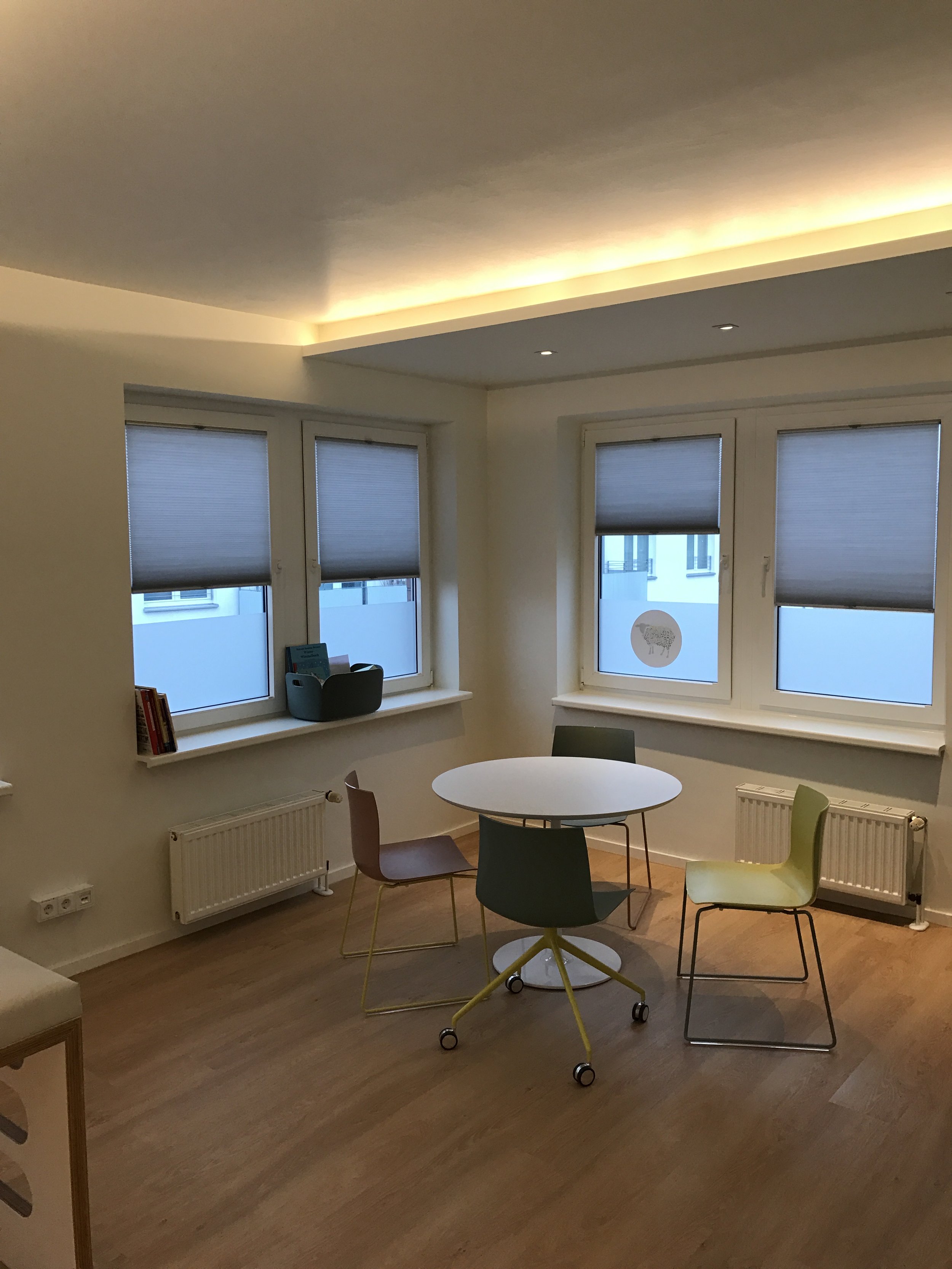 Ein modern eingerichteter Raum mit vier bunten Stühlen um einen weißen runden Tisch, drei Fenster mit Rollläden, Holzboden und indirekter Deckenbeleuchtung.
