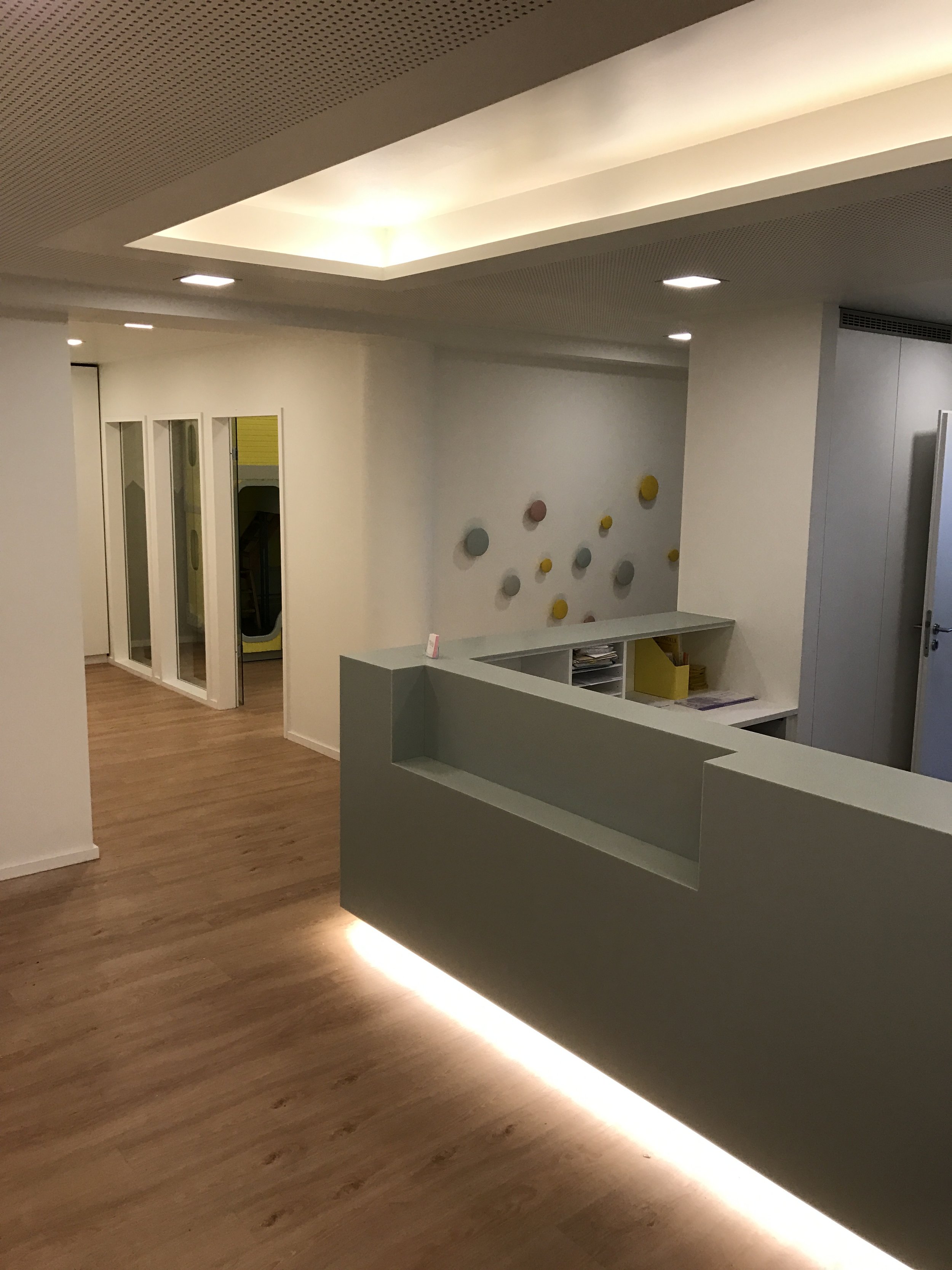 Innenaufnahme eines modernen, hellen Raums mit Holzfußboden, weißer Wanddekoration, und Empfangstresen mit integriertem Licht. Im Hintergrund sind Glasabteile sichtbar.