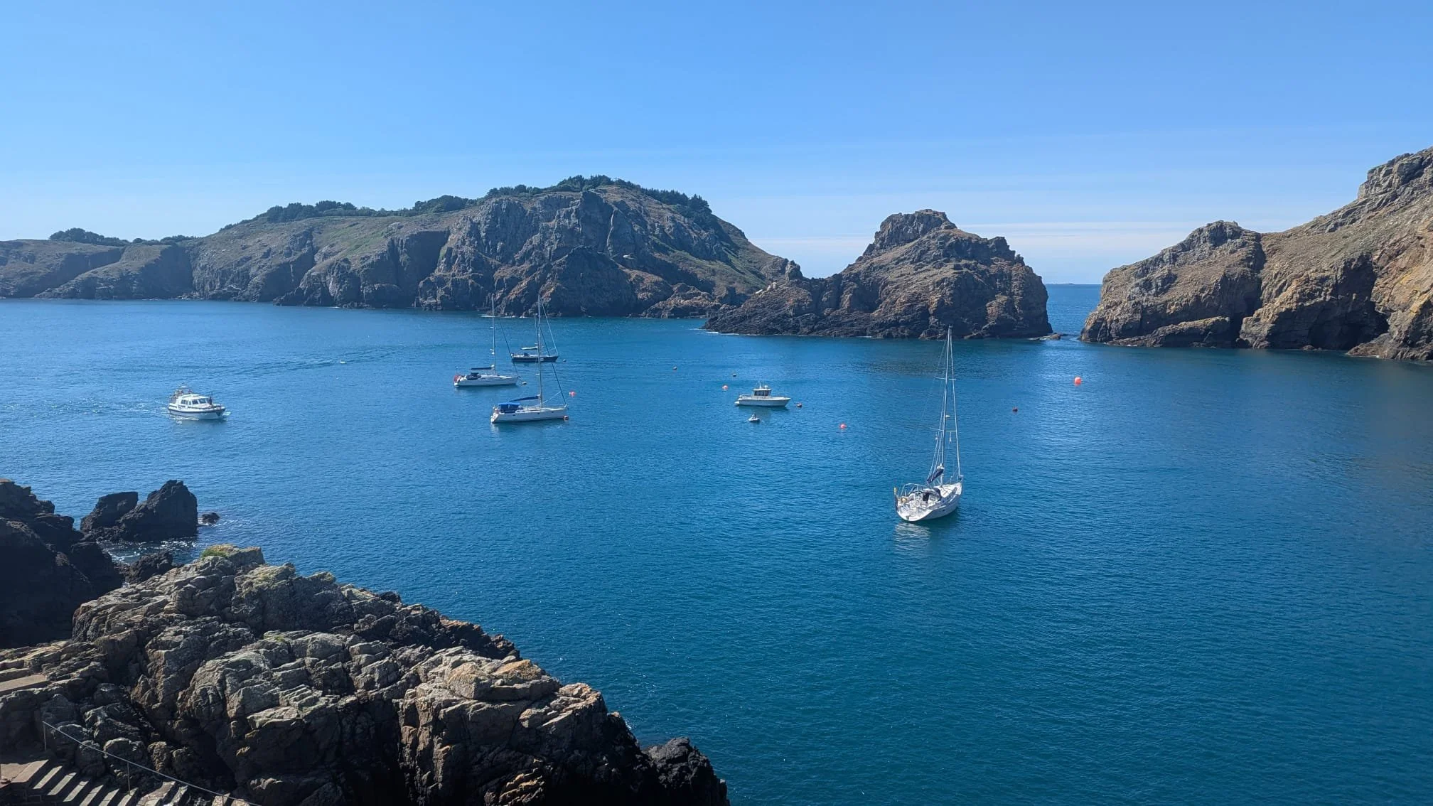 A scenic view of a bay with blue water, several sailboats and yachts, surrounded by rocky cliffs and green hills under a clear blue sky.