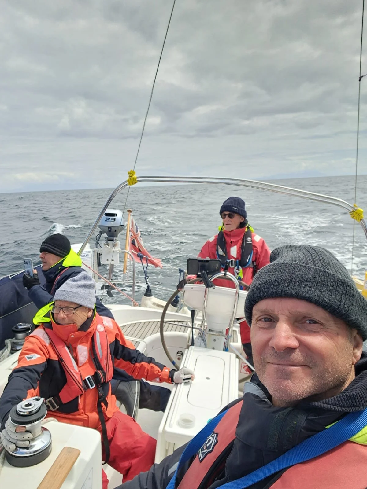 Four people on a boat at sea on a cloudy day. They are wearing life jackets and cold weather clothing, including hats and gloves.
