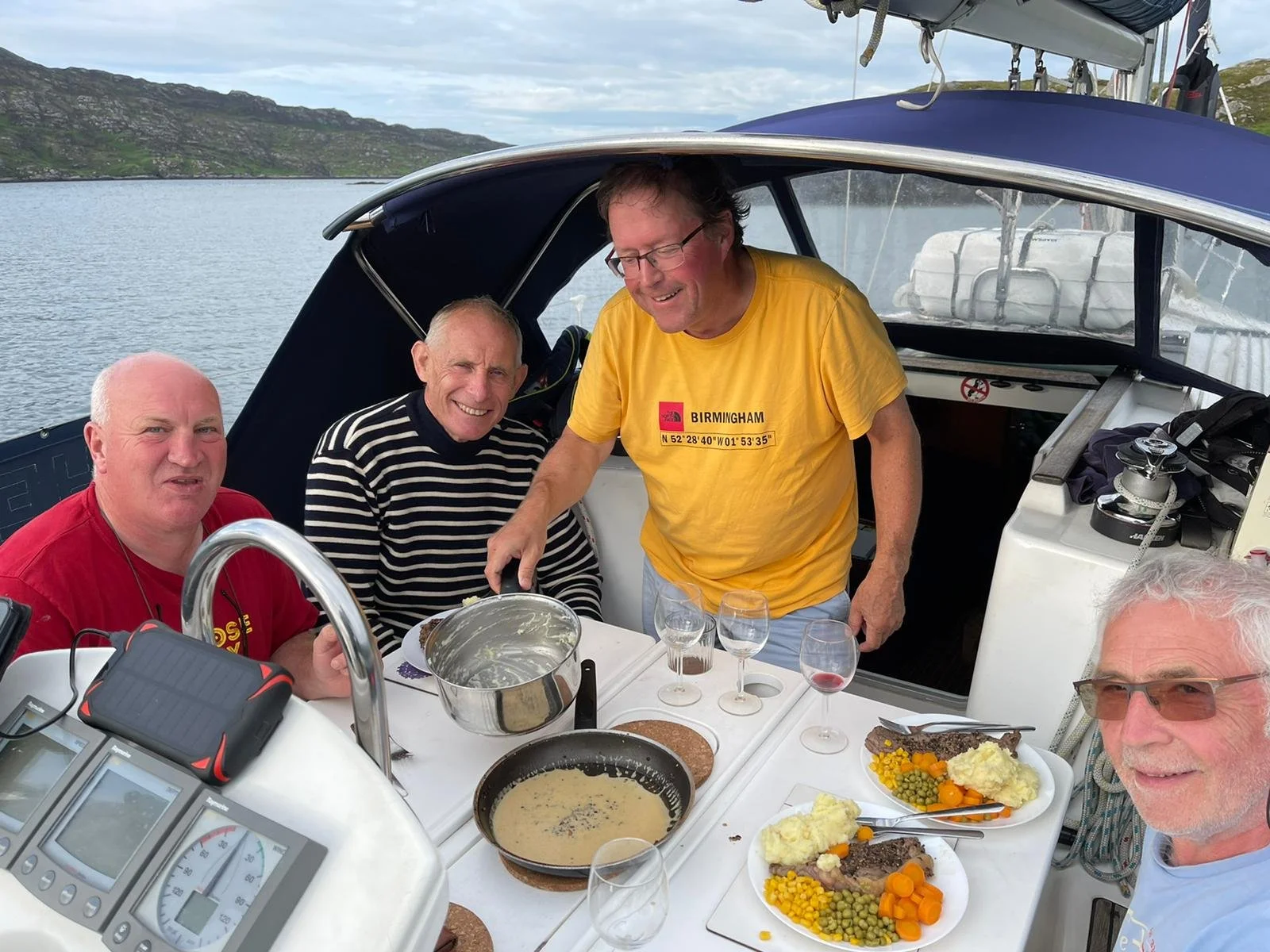 Four men are on a boat enjoying a meal together, with a lake and mountains in the background. They are smiling and there are plates of food with mashed potatoes, vegetables, and meat on the table.