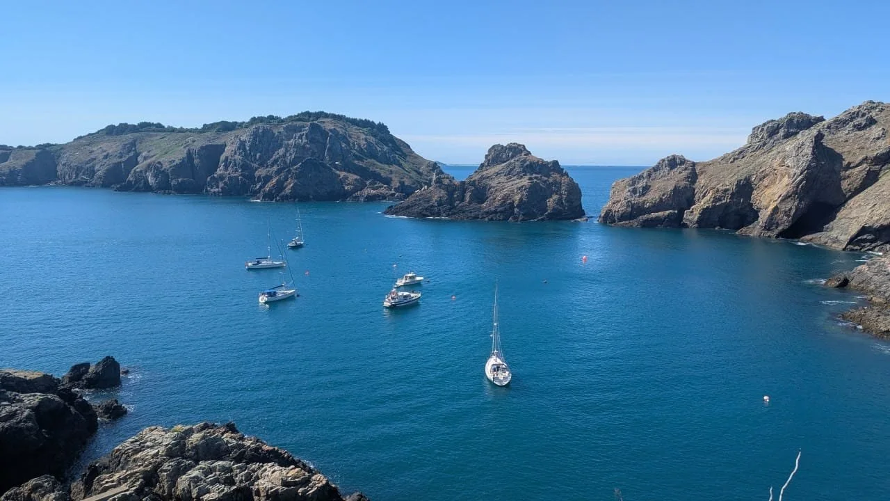 A scenic view of a cove with several boats anchored in calm blue water, surrounded by rocky cliffs and green hills under a clear blue sky.
