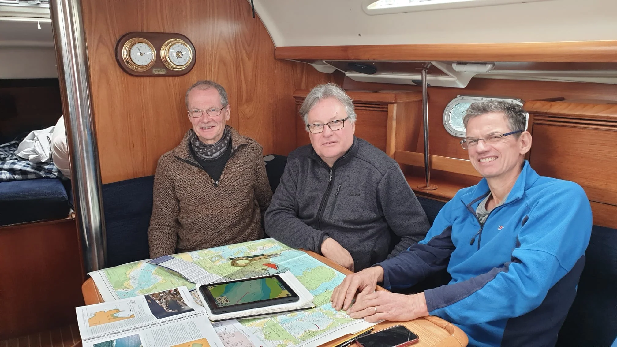 Three men sitting around a table on a boat, with nautical charts, tablets, and magazines on the table, wooden interior, porthole window, and nautical gauges visible on the wall.