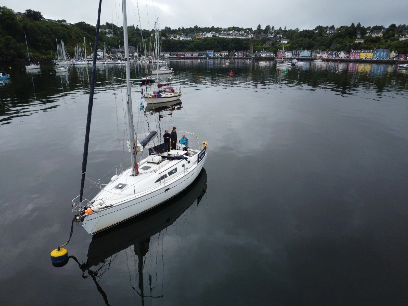 A sailboat with three people on board floating on calm water in a harbor, with colorful houses on a hillside in the background.