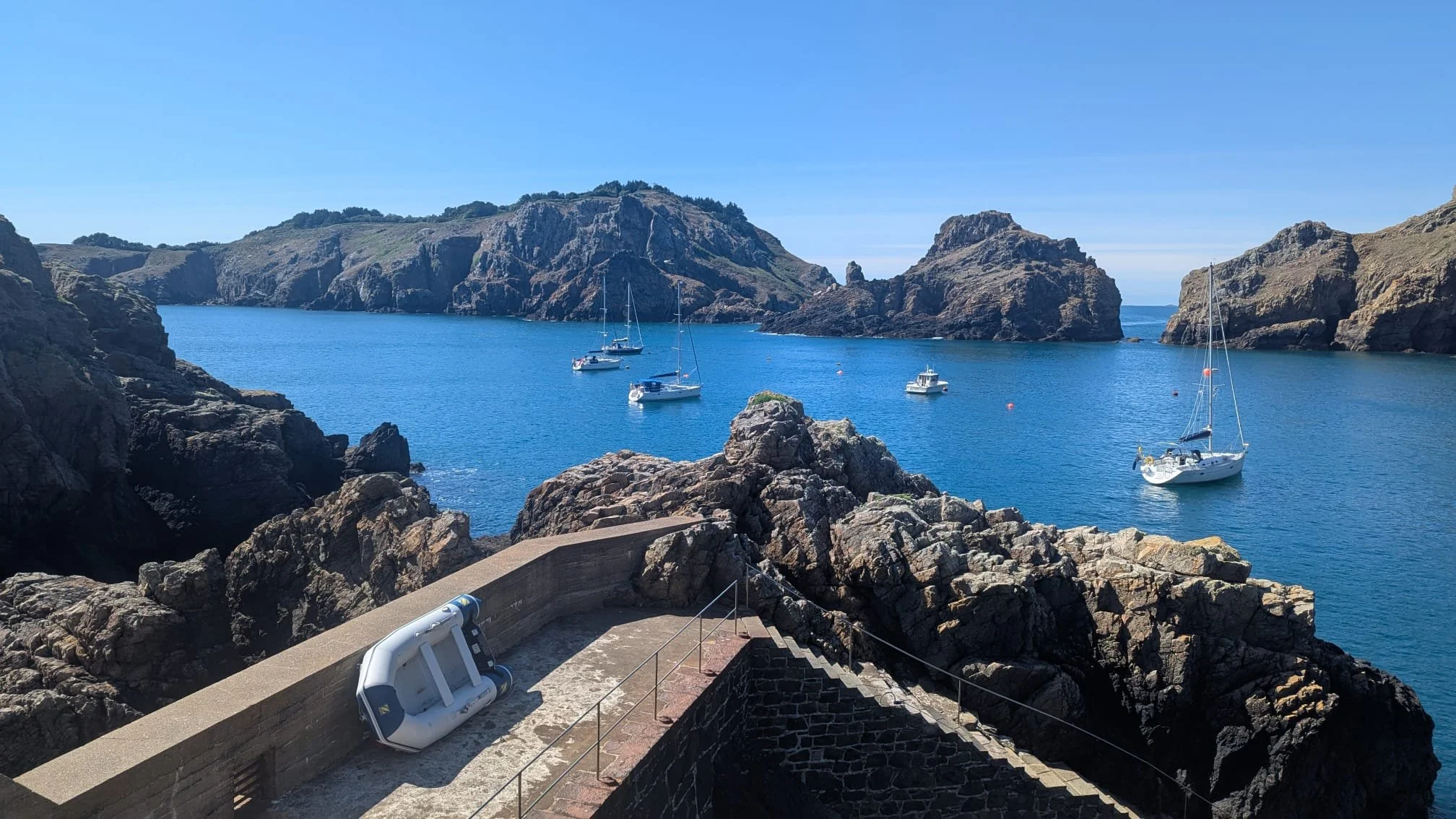 Calm blue sea with five white sailboats anchored near rocky cliffs and hills under a clear blue sky.
