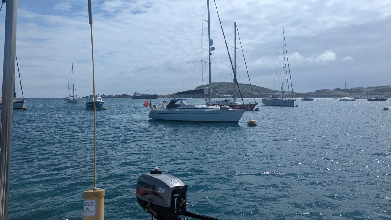 A harbor with several sailboats anchored in calm water, with a hilly landscape and overcast sky in the background. The photo is taken from a boat, as an outboard motor and part of the boat's railing are visible in the foreground.