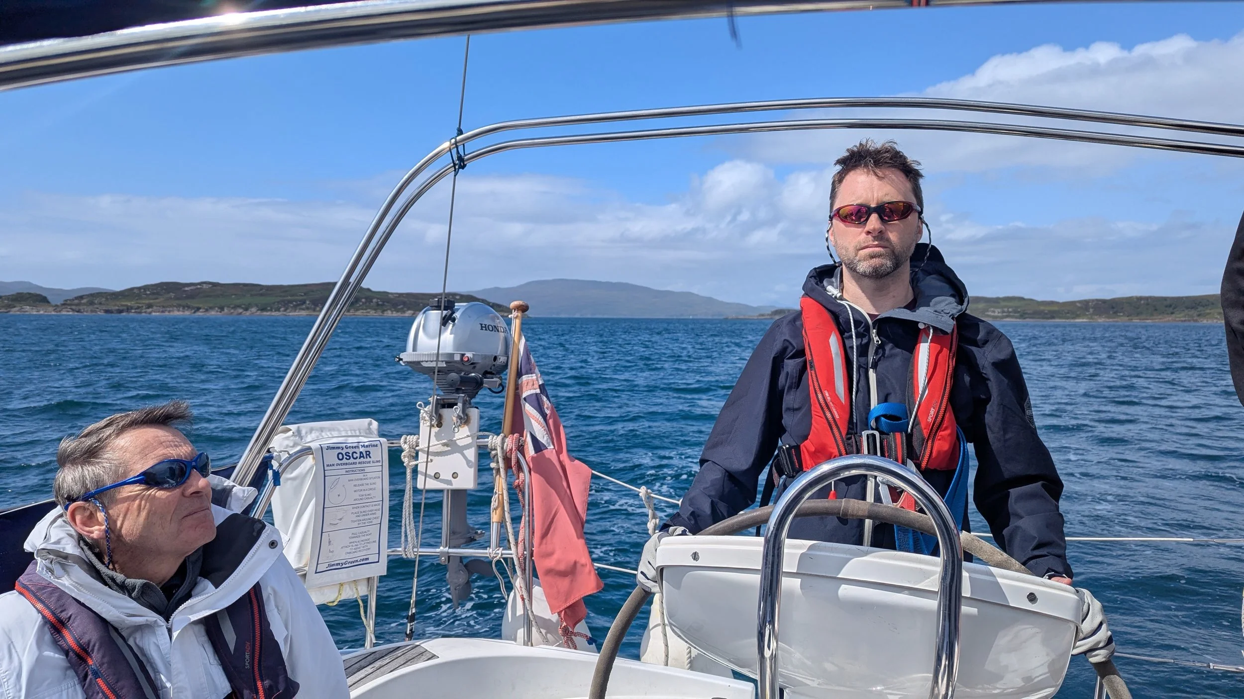 Two men sailing on a boat in the ocean. One is standing at the helm wearing a red life jacket and sunglasses, and the other is sitting wearing sunglasses and a white jacket. There are hills in the background under a partly cloudy sky.