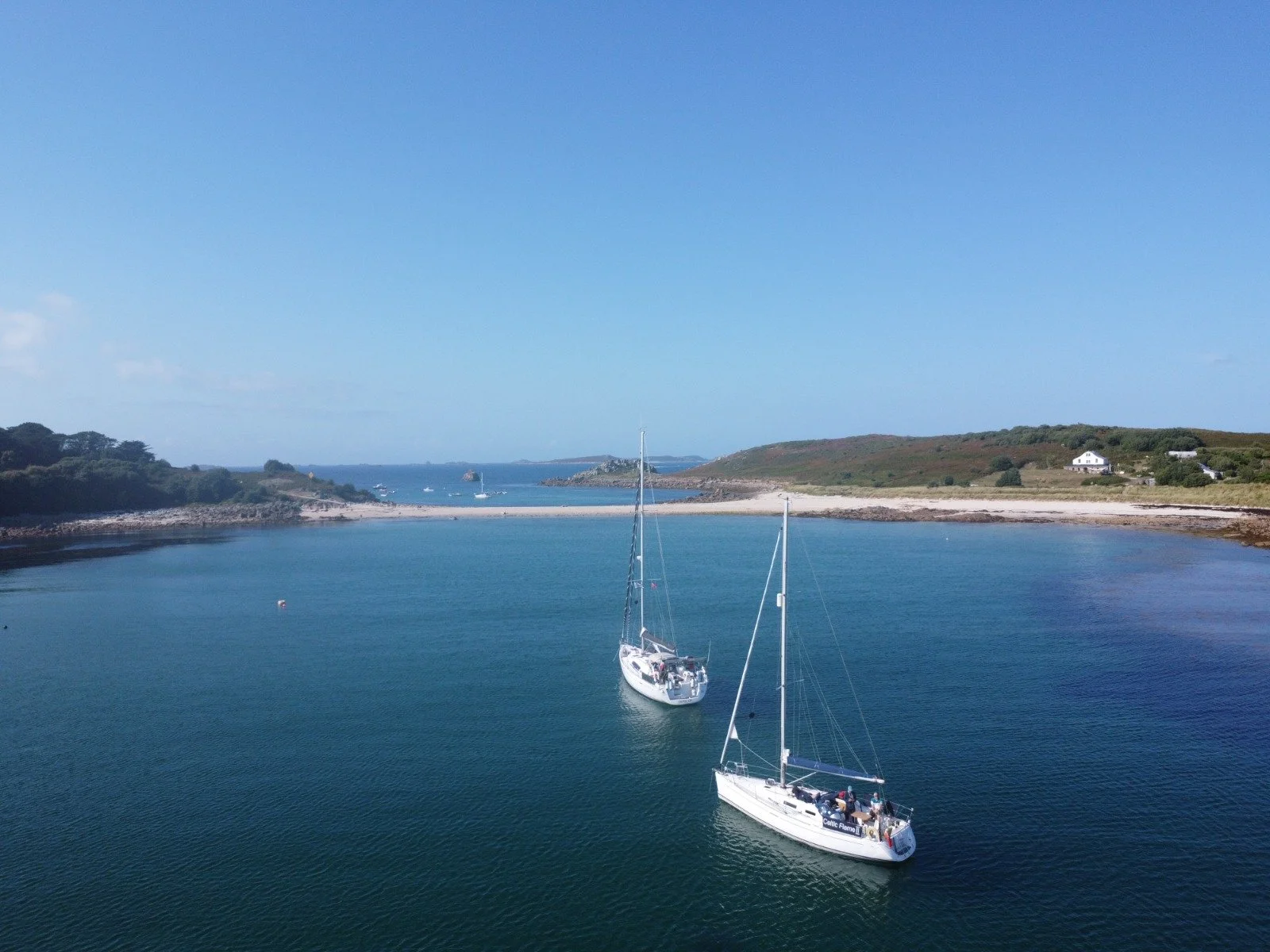Two sailboats in a calm, blue bay with a sandy shore, green hills, and distant houses under a clear sky.