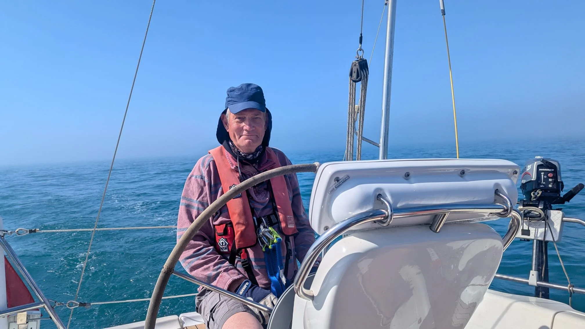 A man sailing on a boat in the open ocean, wearing a cap, a life jacket, and a long-sleeve shirt, with a calm blue sea and sky in the background.