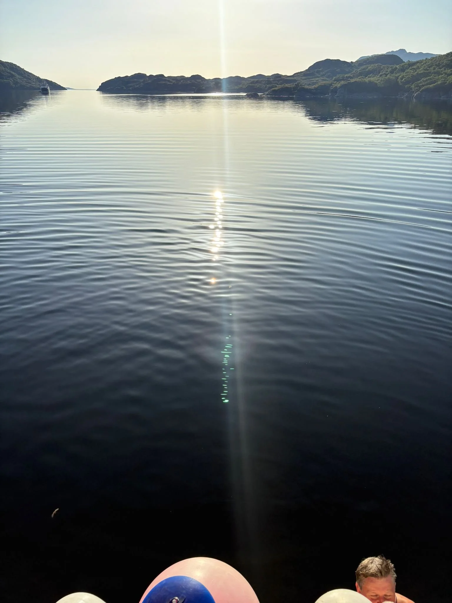 A body of water reflecting the sky with small waves, surrounded by hills and mountains, with a boat in the distance and a person's legs and face visible at the bottom of the frame.