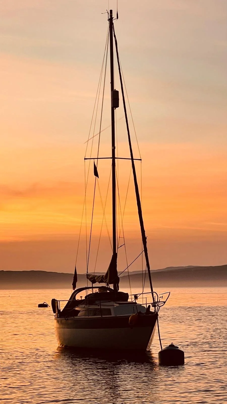 Sailboat anchored on calm water during a colorful sunset with a distant shoreline in the background.