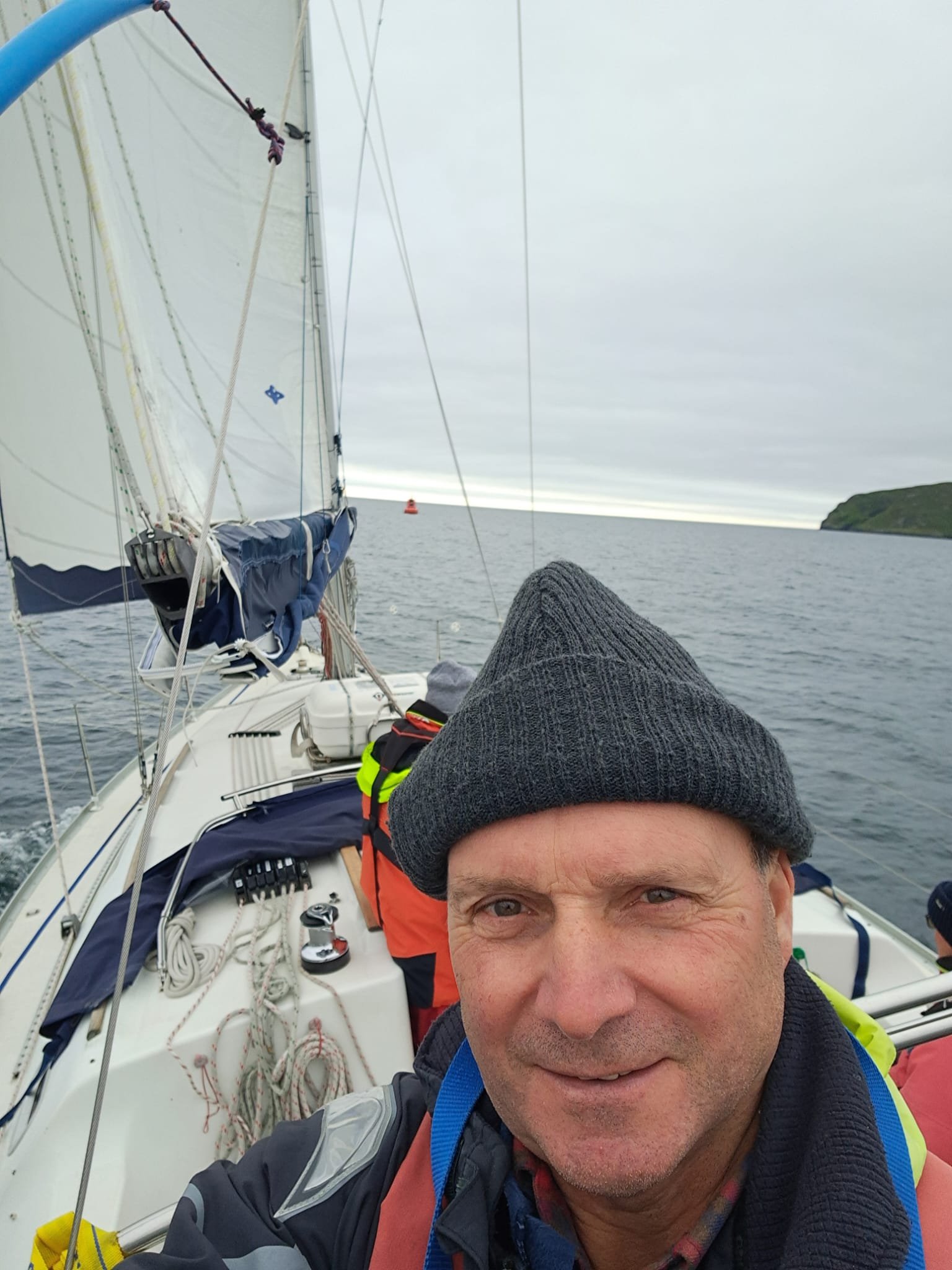A man taking a selfie on a sailboat with gray skies and water in the background, wearing a gray beanie and a colorful jacket.