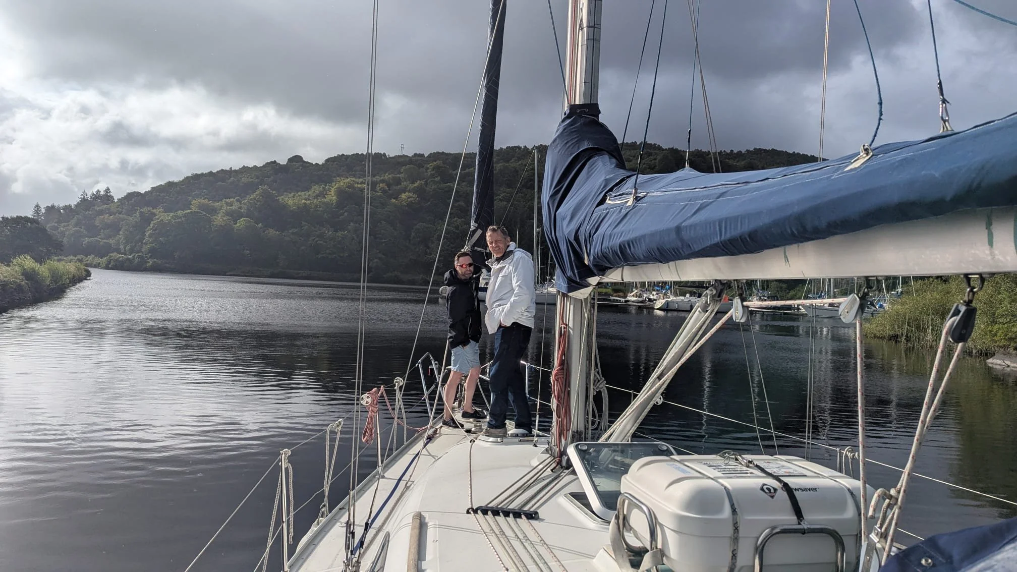 Two men standing on a sailboat deck, with water and green hills in the background, under cloudy skies.
