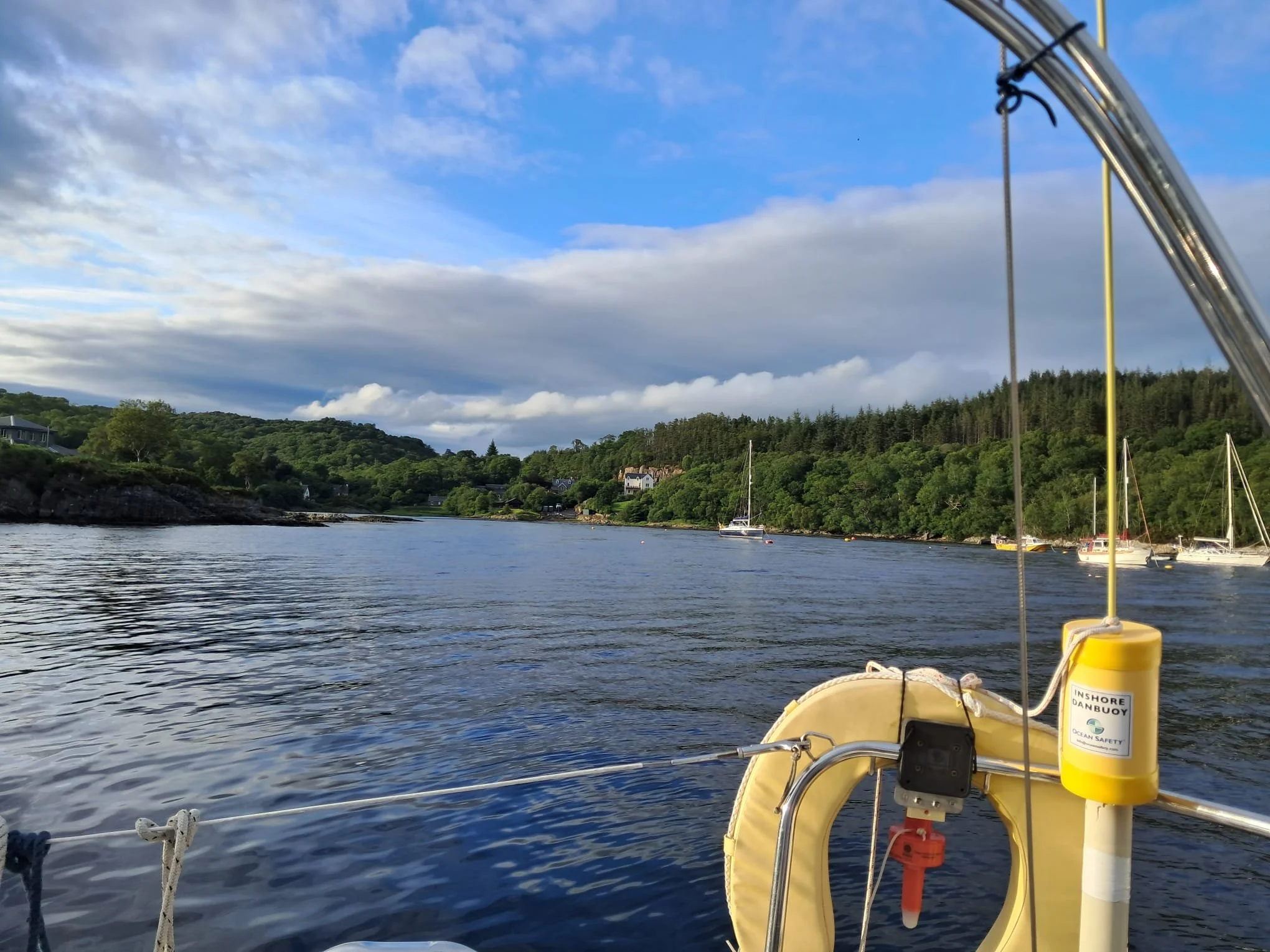View from a boat on a river, with a life buoy and safety equipment in the foreground, lush green trees on the riverbank, and sailboats moored along the shore under a partly cloudy sky.
