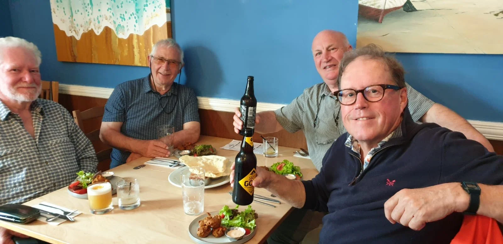 Four men sitting at a dining table with food and drinks, smiling for a photo during a gathering or celebration.