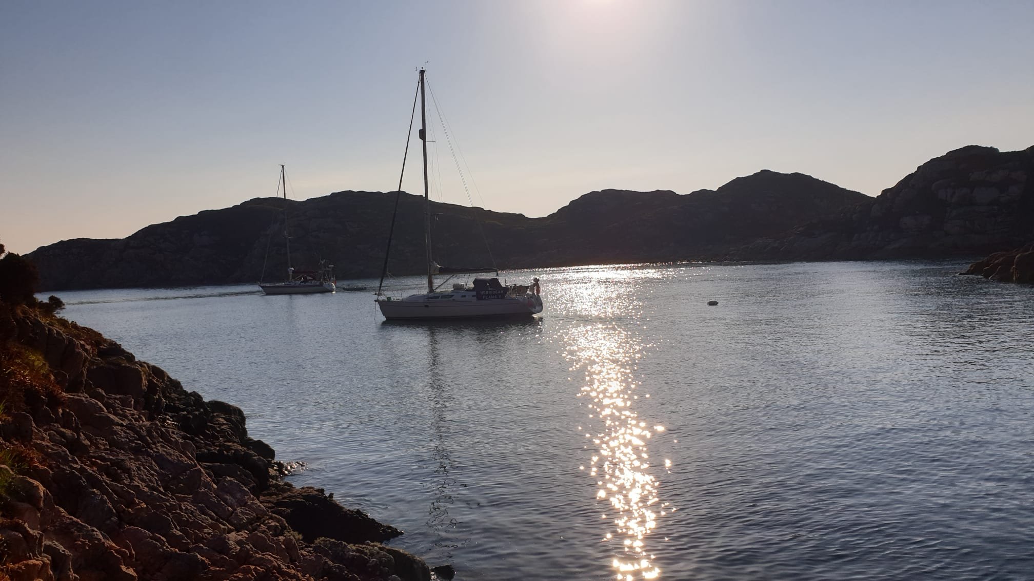 A calm body of water with two sailboats anchored, surrounded by rocky terrain and hills in the background, with the sun reflecting on the water's surface.