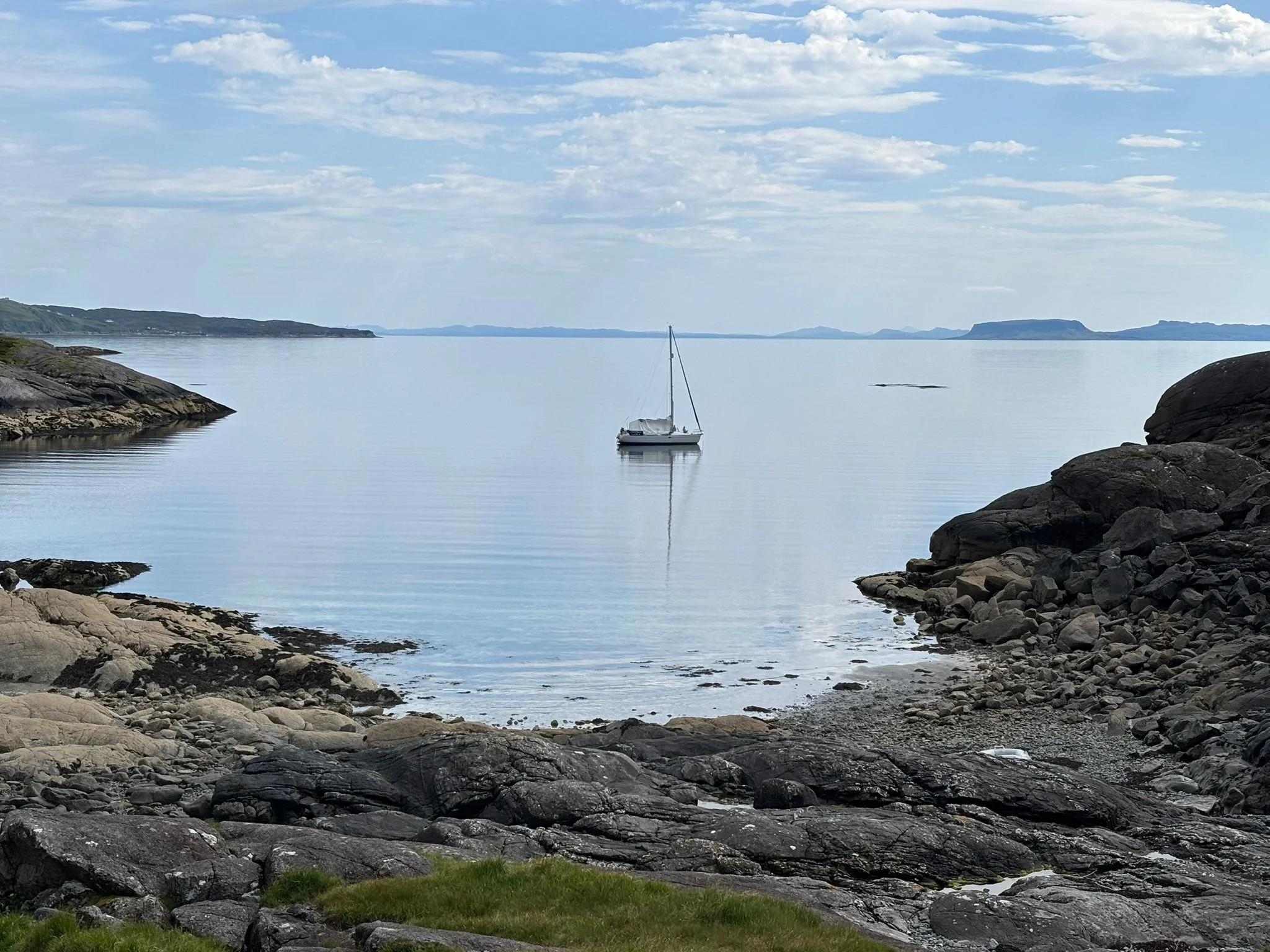 A calm sea with a single sailboat anchored near rocky shores under a partly cloudy sky.
