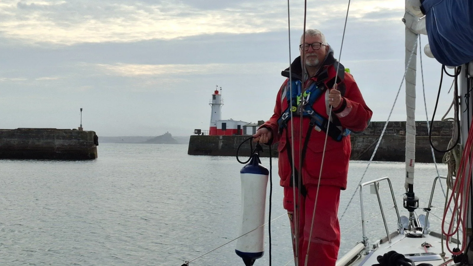 A man in red sailing gear standing on a sailboat, holding a fender with water in the background and a lighthouse along with rocks and the ocean visible in the distance during cloudy weather.