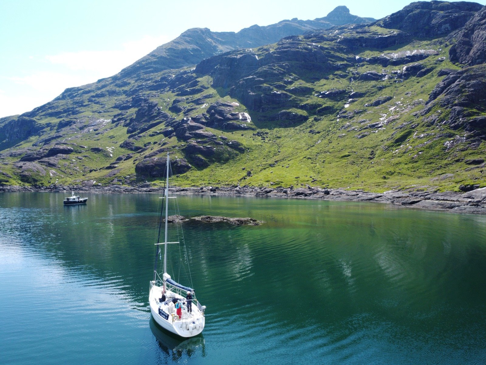 Sailboats on a calm body of water with green mountains and rocky terrain in the background.