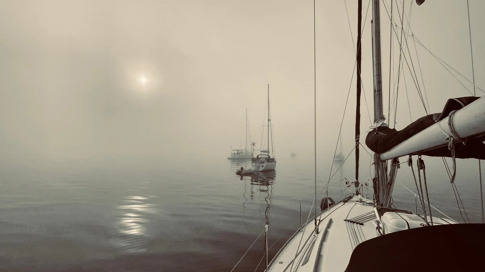 A foggy scene on the water with sailboats anchored and a person in the distance near one of the boats.