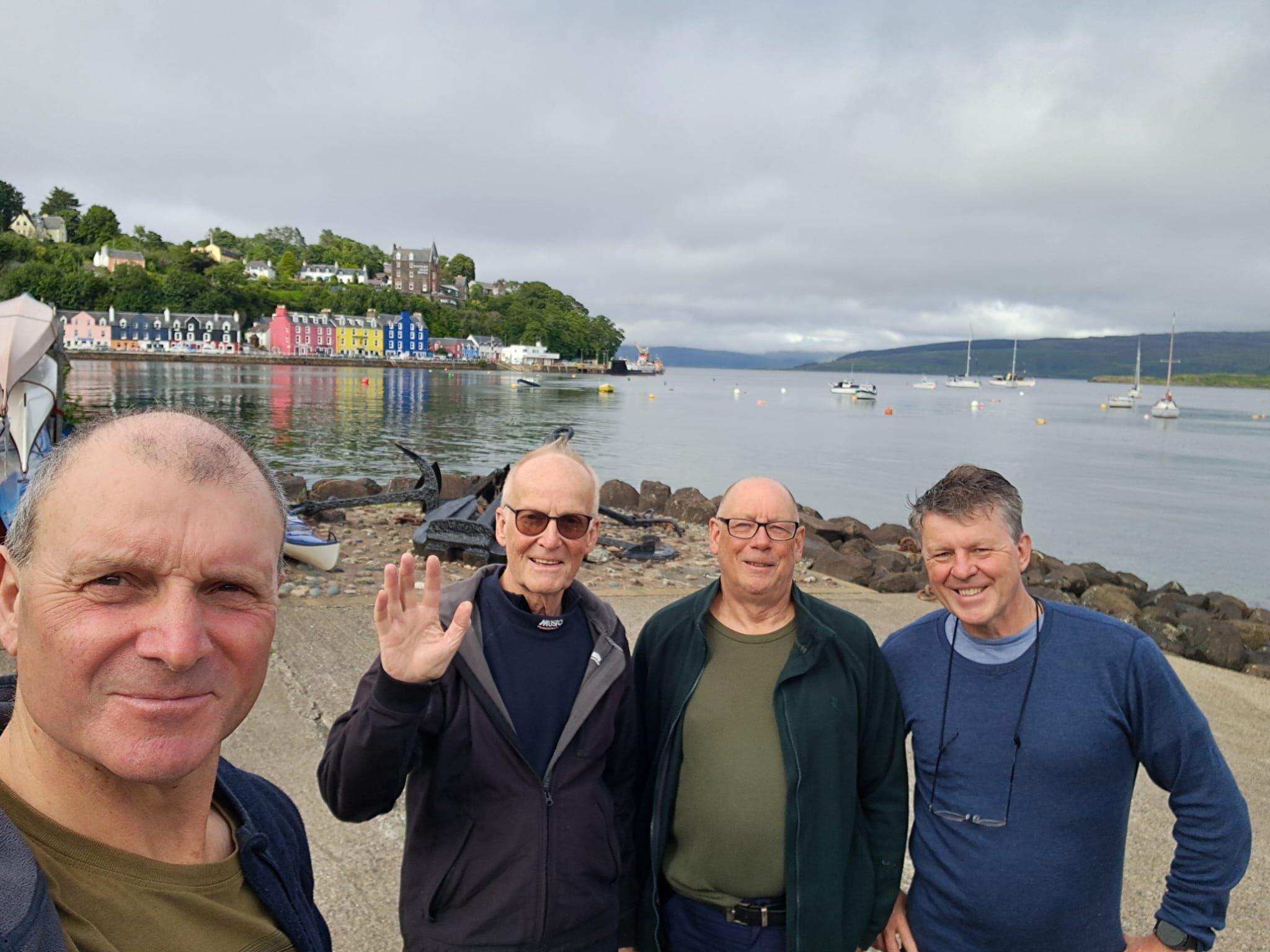 Four men standing on a waterfront in front of colorful houses and boats with rocky shoreline and cloudy sky.