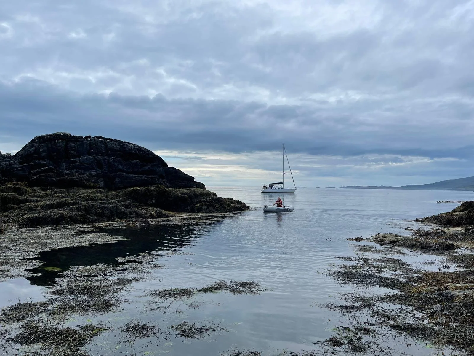 A calm coastal scene with rocky shoreline and two boats, one small and one larger, floating on the water under a cloudy sky.