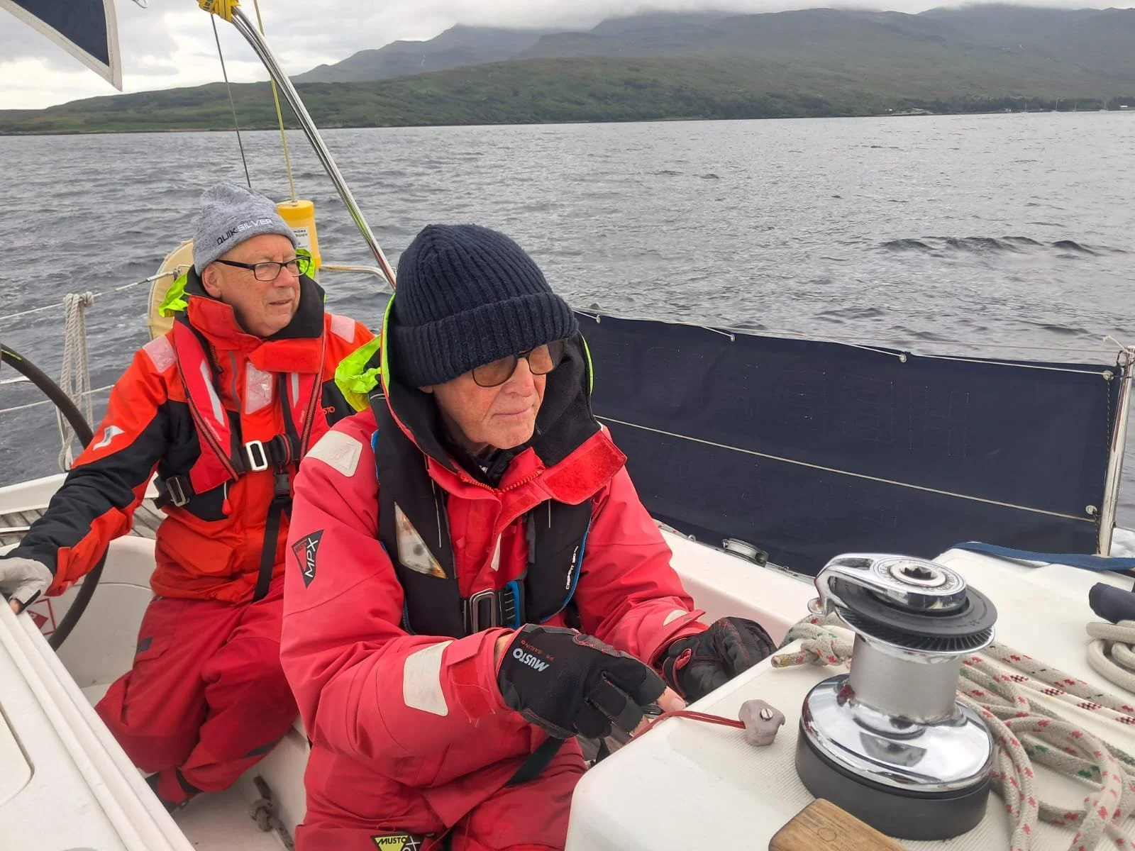 Two older men dressed in red waterproof jackets and winter hats aboard a sailboat, with a body of water and mountains in the background.