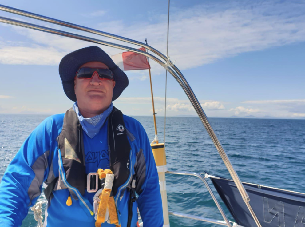 A man wearing a blue jacket, hat, and sunglasses standing on a boat in the ocean under a partly cloudy sky.