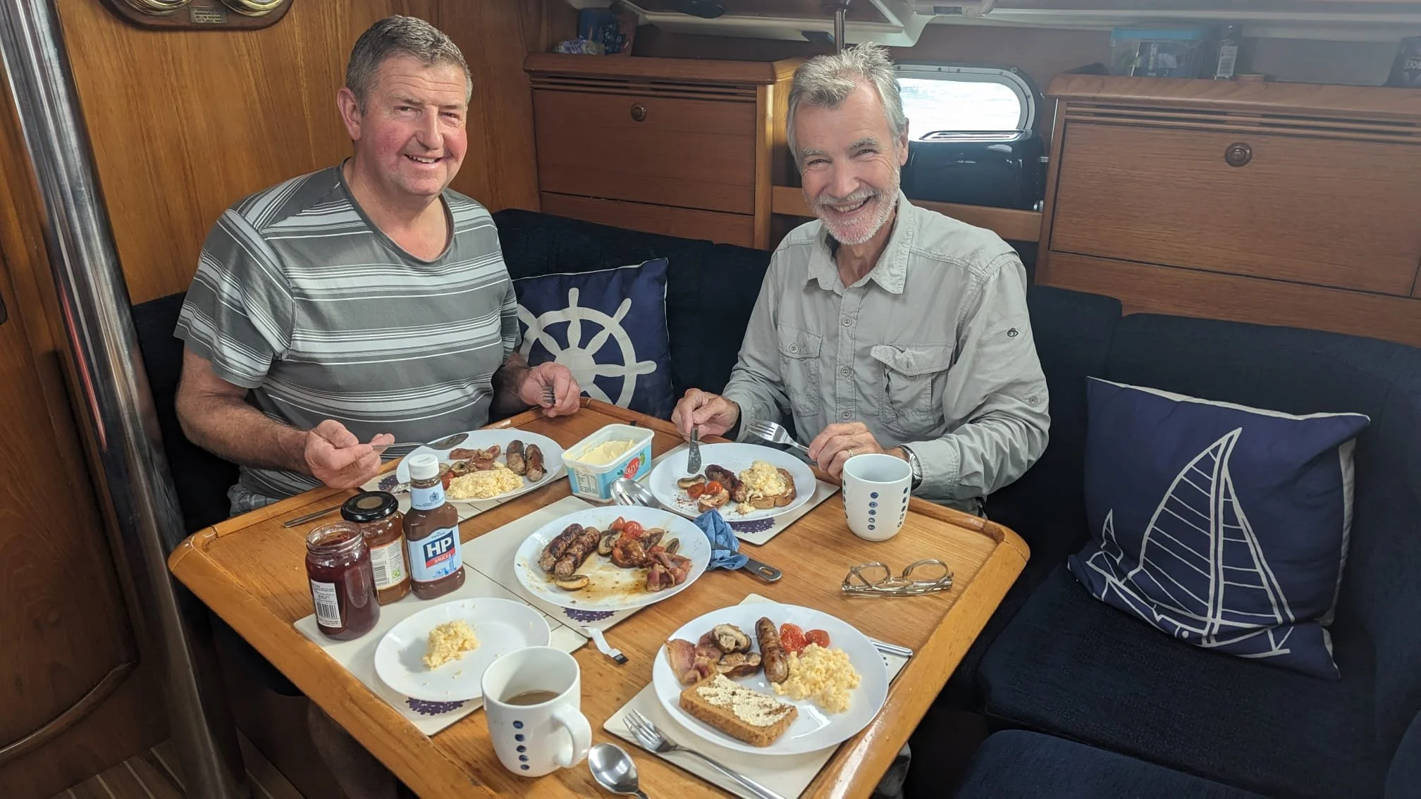 Two men sitting at a table enjoying breakfast inside a boat. The table has plates with sausages, bacon, eggs, toast, and tomato; there are also cups, jars of jam, butter, and condiments. The men are smiling and holding utensils, with nautical-themed 