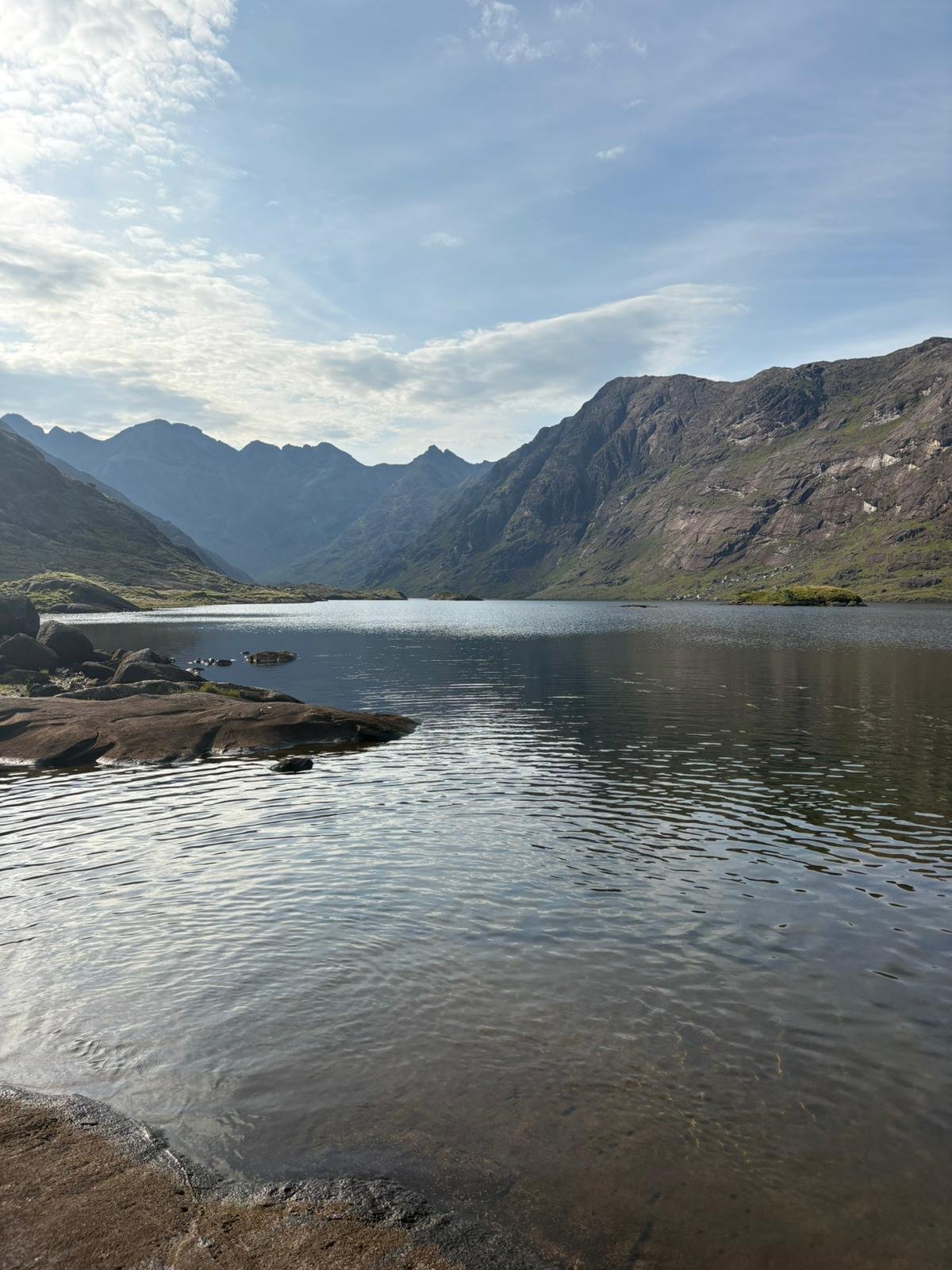 Scenic view of a mountain lake with rocky shoreline, surrounded by tall rugged mountains under a partly cloudy sky.
