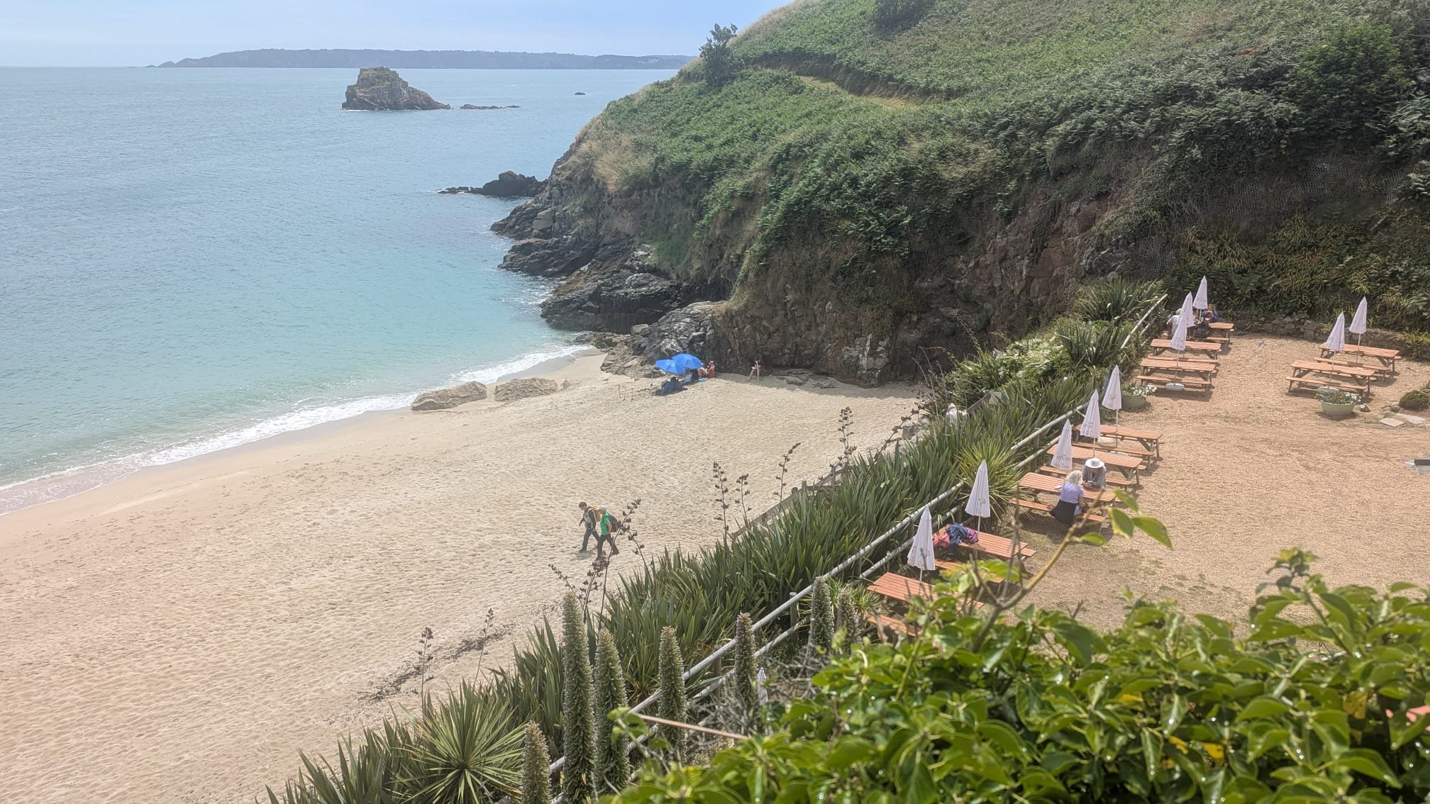 A beach with light-colored sand, ocean water, rocky cliffs, and green vegetation. There are a few people under an umbrella and on the sand, with empty lounge chairs and umbrellas nearby.