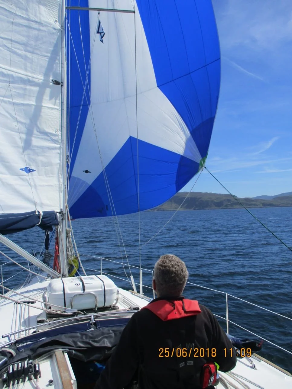 A person with short hair wearing a black jacket with a red collar is sitting on a sailboat, navigating with blue and white spinnaker sails in the open water under a clear blue sky, with hills in the background.