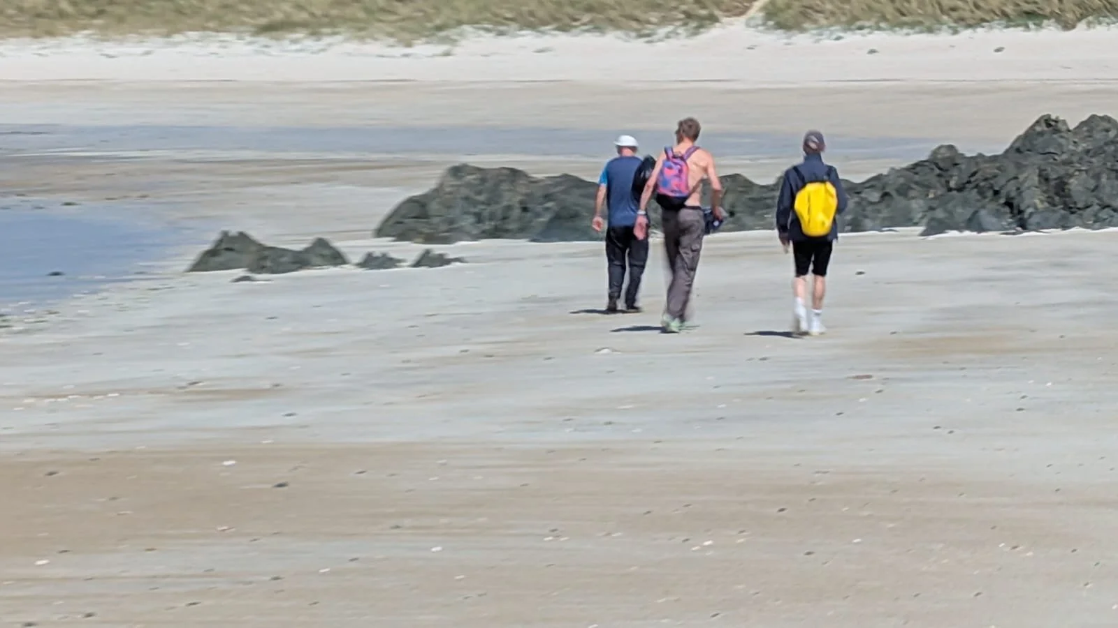 Three people walking on a beach, one with a pink backpack, one with a yellow backpack, and one with no visible pack, near rocks and sand.