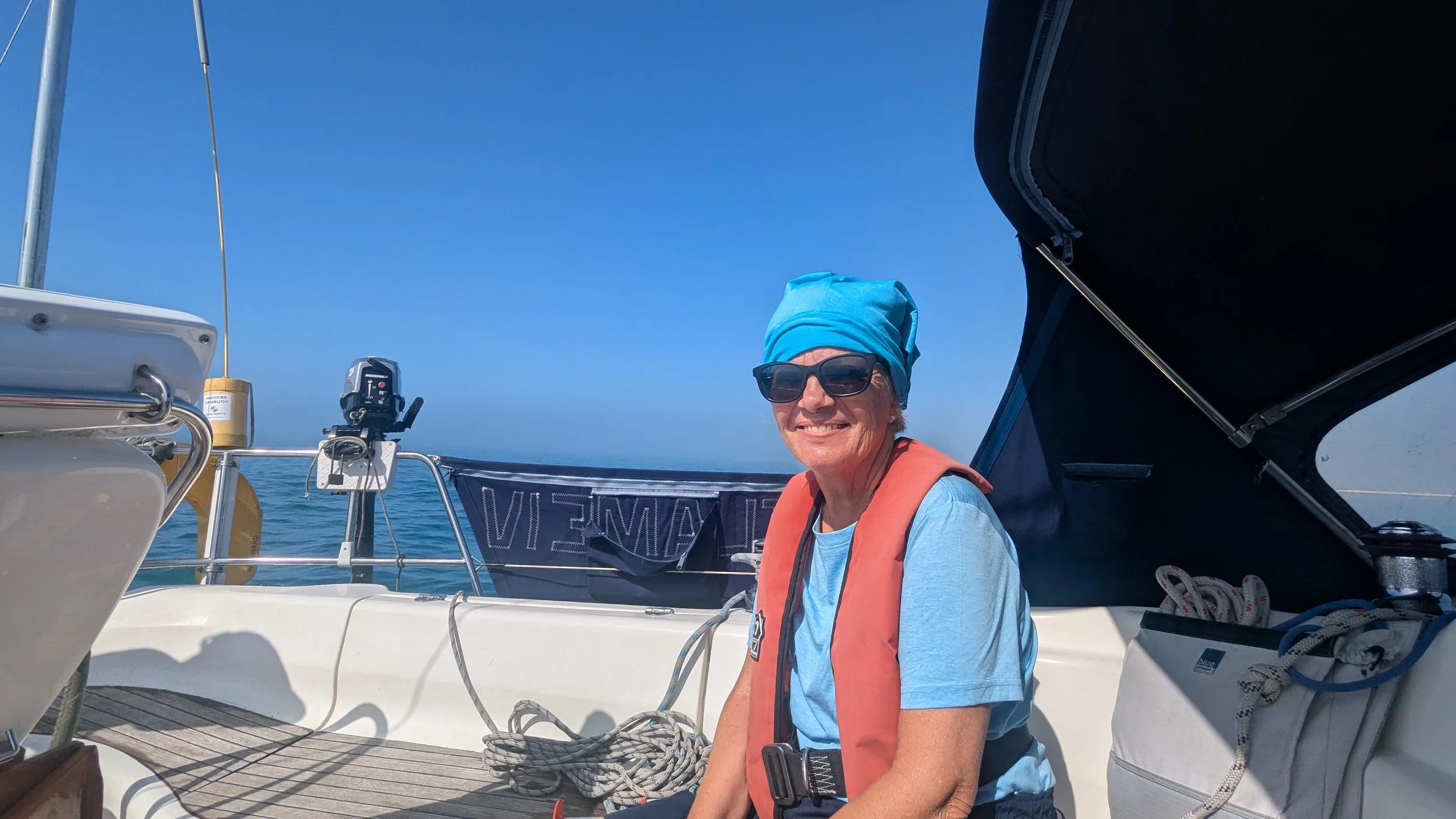 Smiling woman with sunglasses, blue headscarf, and orange life jacket sitting on a boat under a clear blue sky, with sailing equipment and ocean in the background.