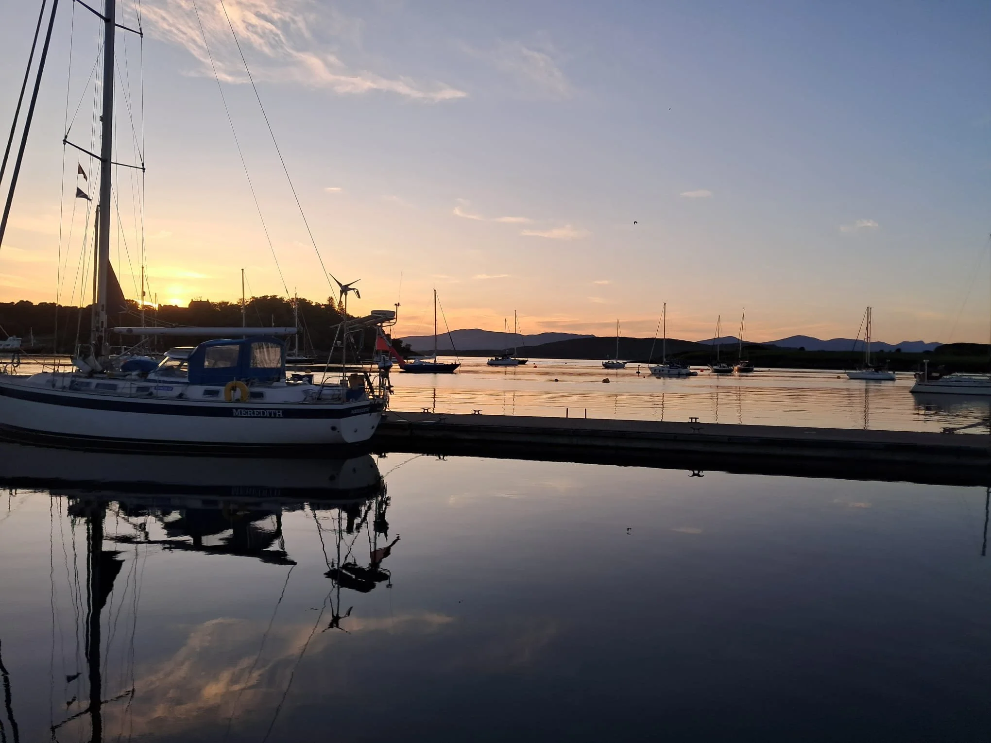 A calm marina at sunset with several sailboats docked, including one named Meredith in the foreground. The sky has soft colors with a few clouds, and the water reflects the boats and the sky. In the background, hills are visible.