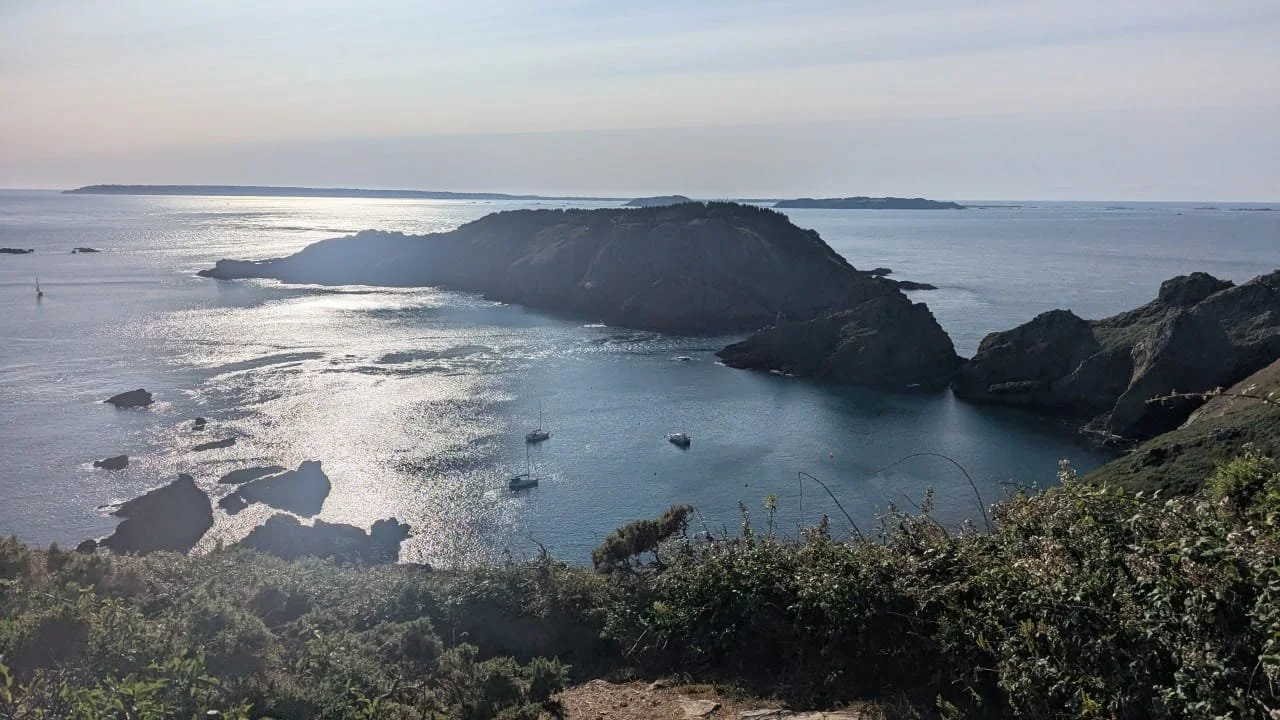 A scenic view of a coastal cove with rocky cliffs, several sailboats floating in calm water, and a distant horizon under a cloudy sky.