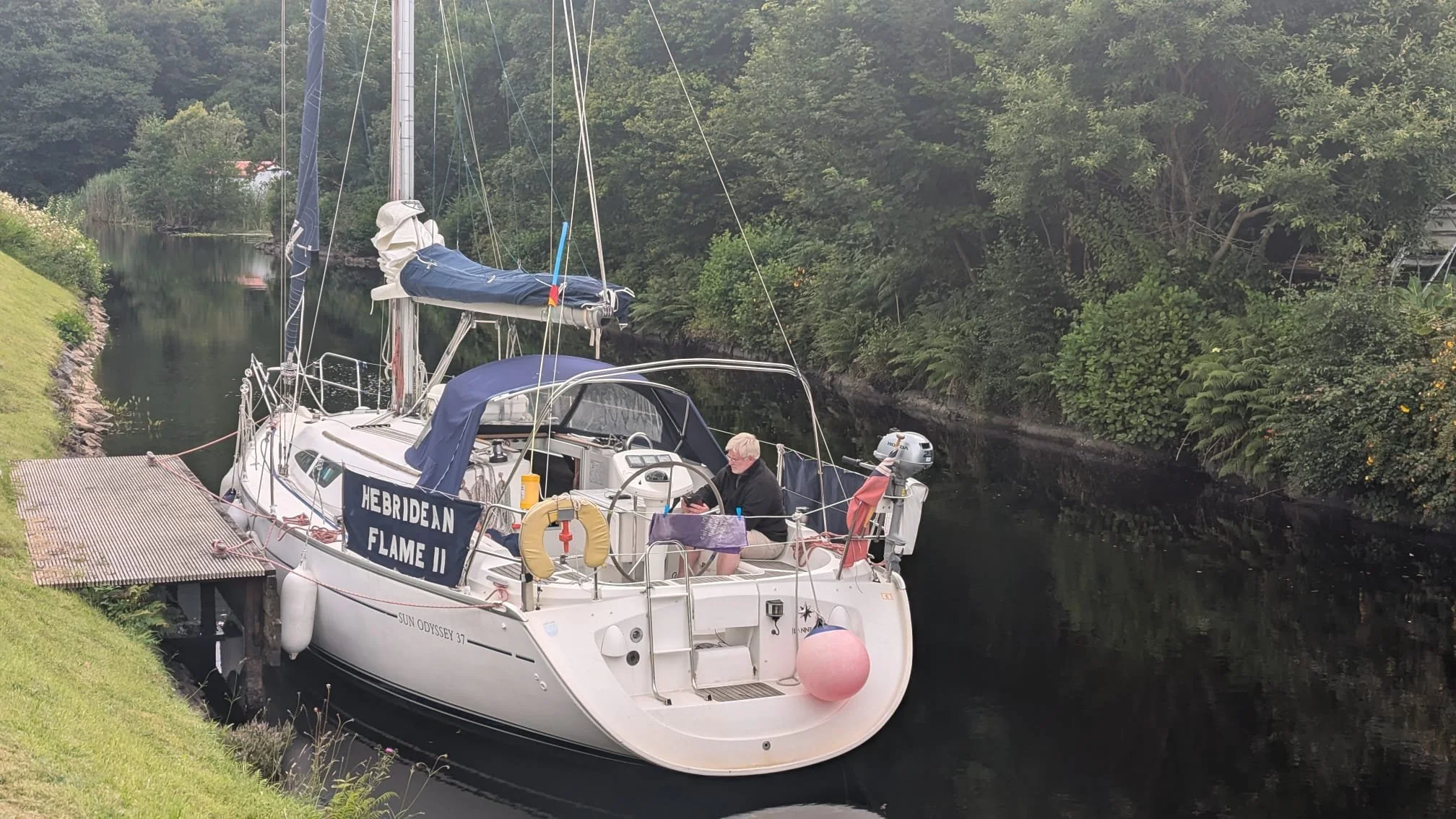 A sailboat docked in a calm canal surrounded by trees, with a person sitting on the deck looking at their phone.