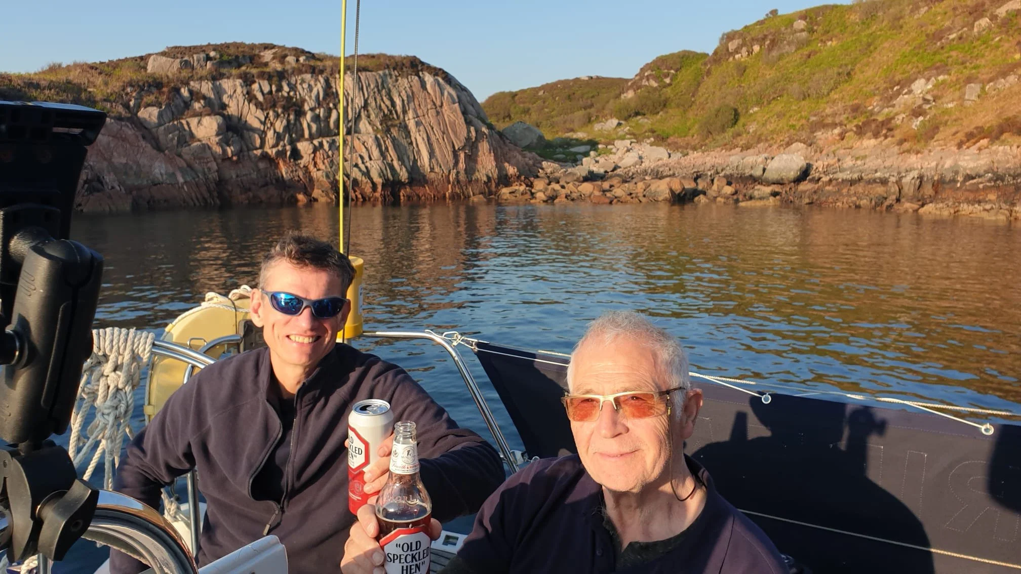 Two men on a boat near rocky shoreline, holding drinks, one wearing sunglasses and smiling, the other wearing tinted glasses, with water and hills in the background.