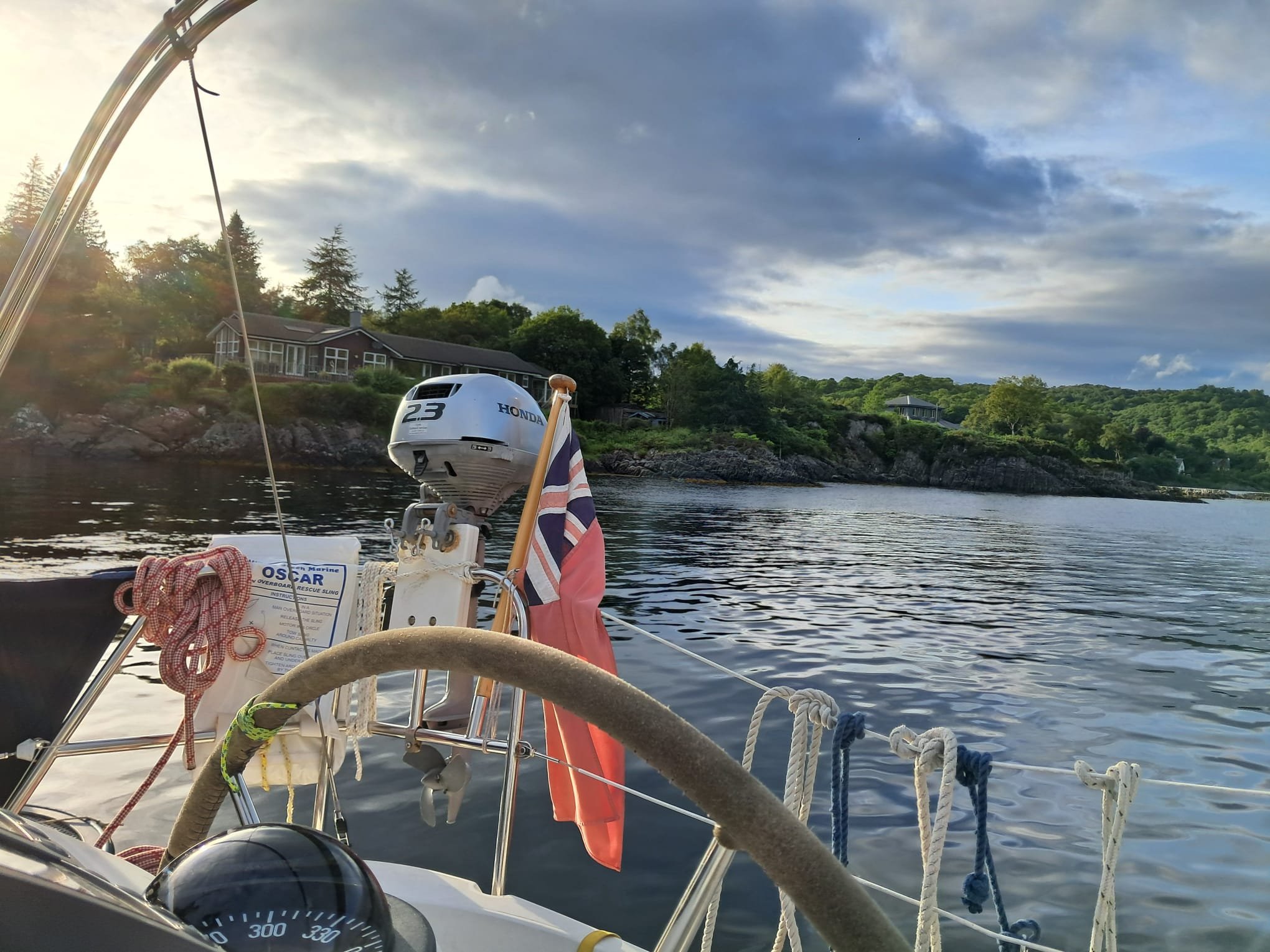 View from a boat on calm water with houses and trees on a hillside in the background, cloudy sky, and boating equipment visible in the foreground.