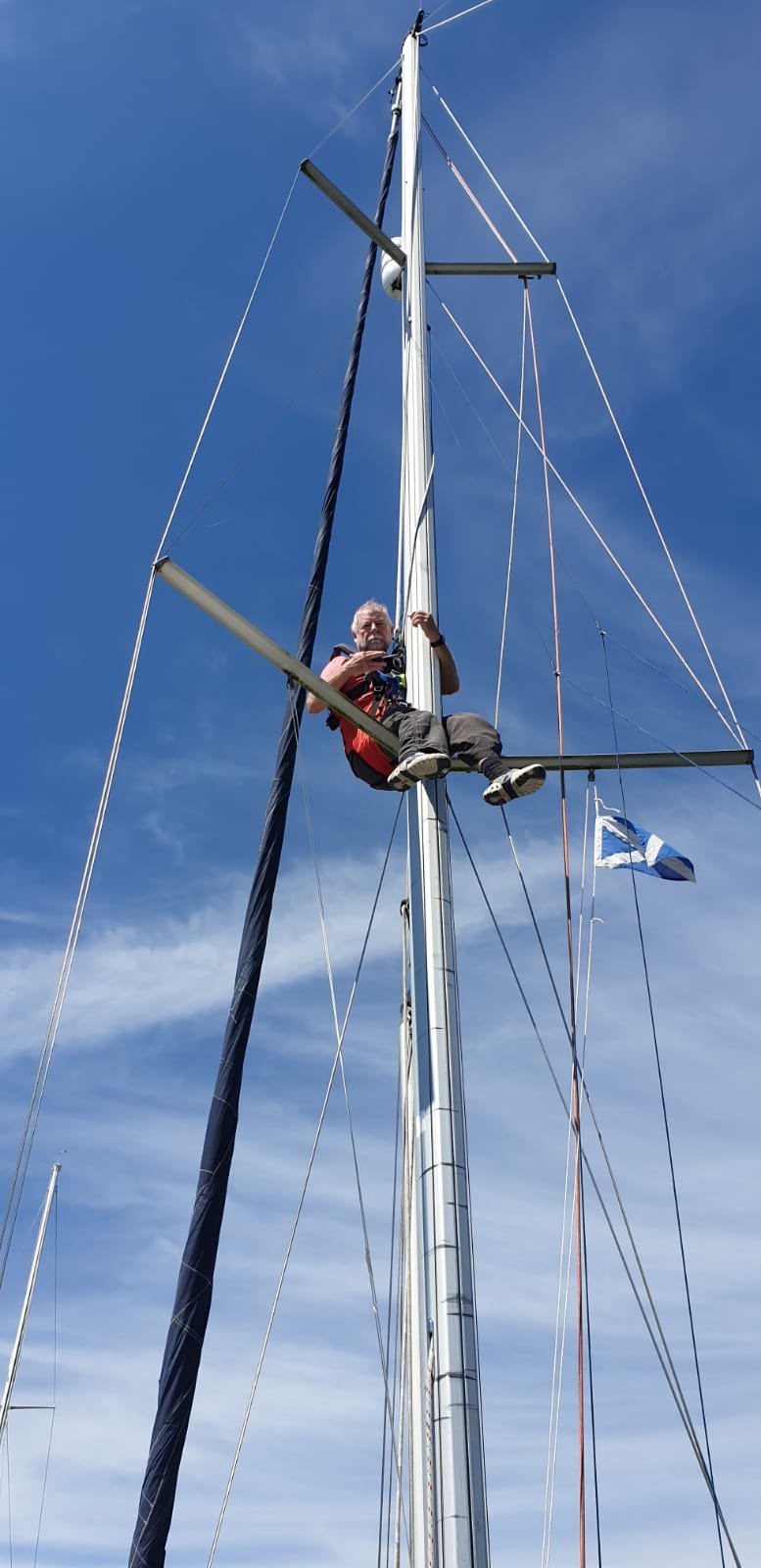 A person climbing or sitting on the horizontal support of a sailboat's mast, with a blue sky and some clouds in the background, and a Finnish flag flying near the base of the mast.