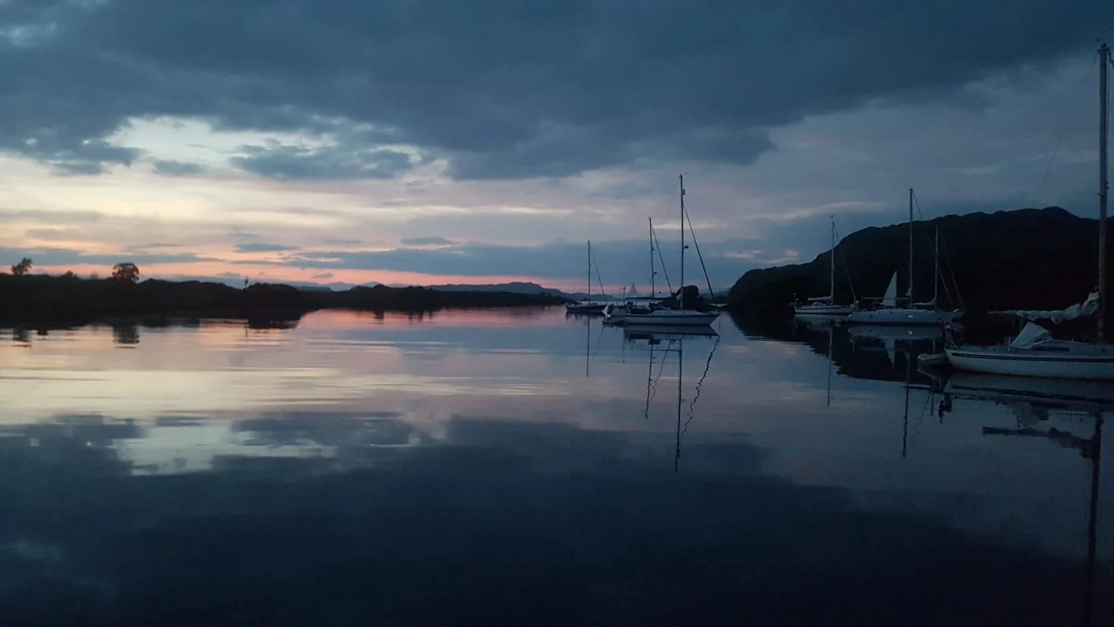 A peaceful scene of sailboats docked at a marina during sunset, with pink and blue hues in the sky and their reflections on the calm water.