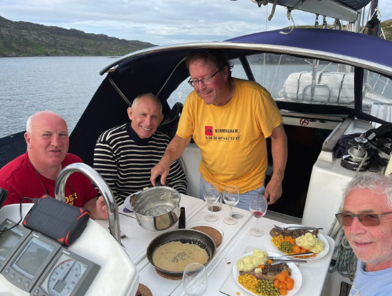 Four men are on a boat having a meal, with a water body and hills in the background. One man is pouring a drink, and the table has food, drinks, and a pan.