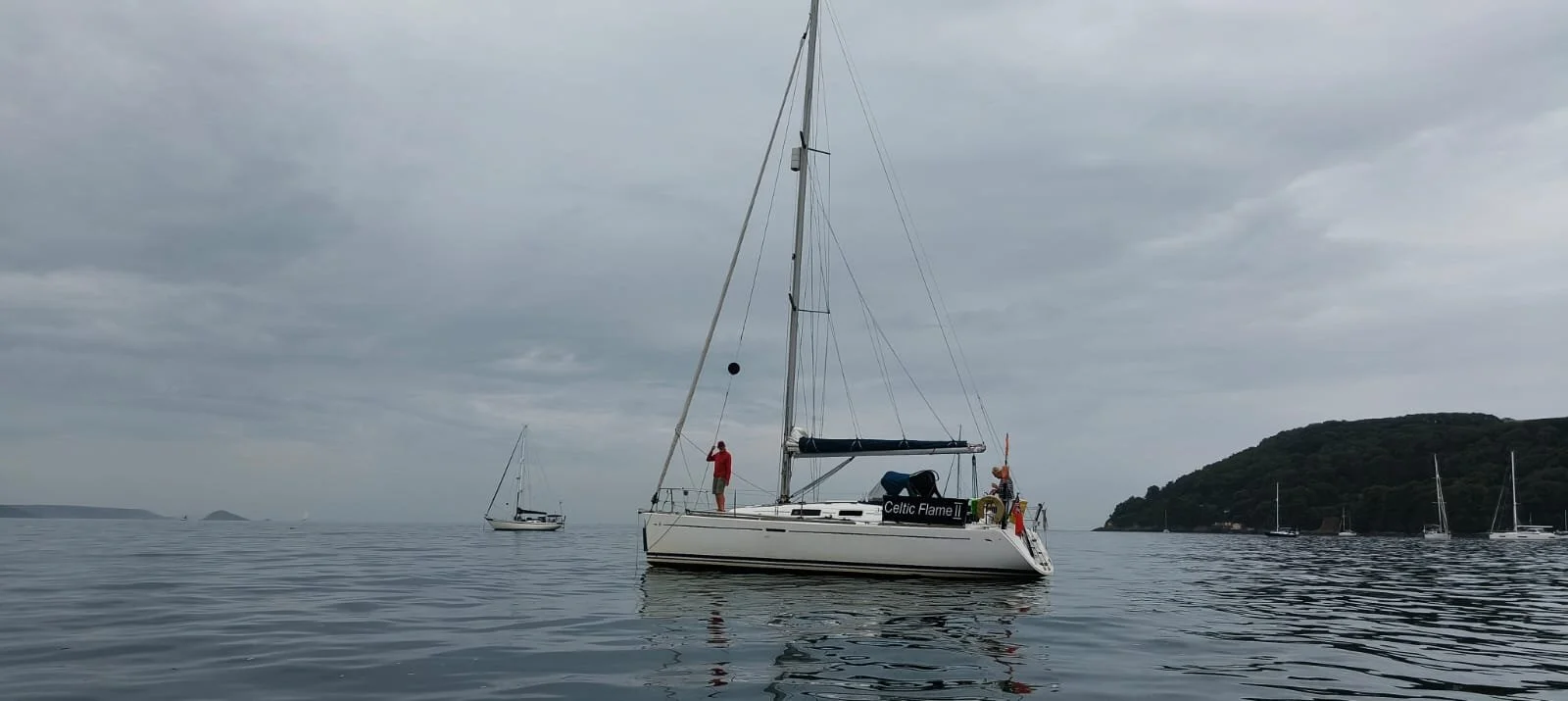 A sailboat on calm water with two people on board, one standing and the other sitting, under a cloudy sky, with other sailboats and a landmass in the background.