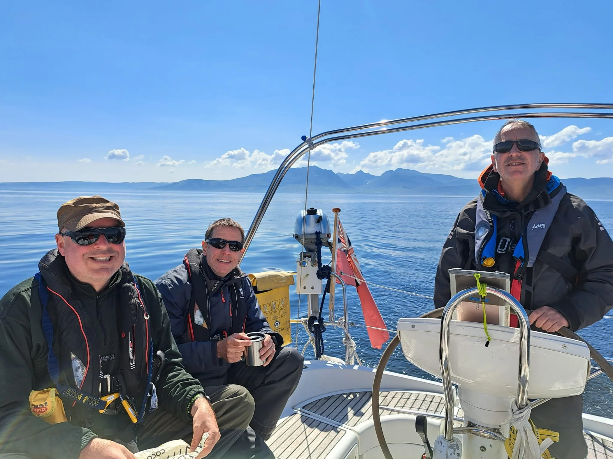 Three men on a boat with a scenic lake and mountains in the background, smiling and wearing outdoor gear and sunglasses.
