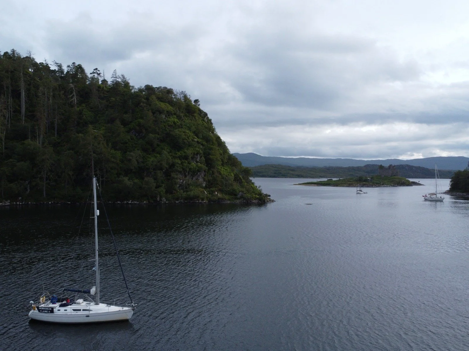 A white sailboat with a tall mast floating on a calm river surrounded by green forested hills and mountains, with a cloudy sky overhead.