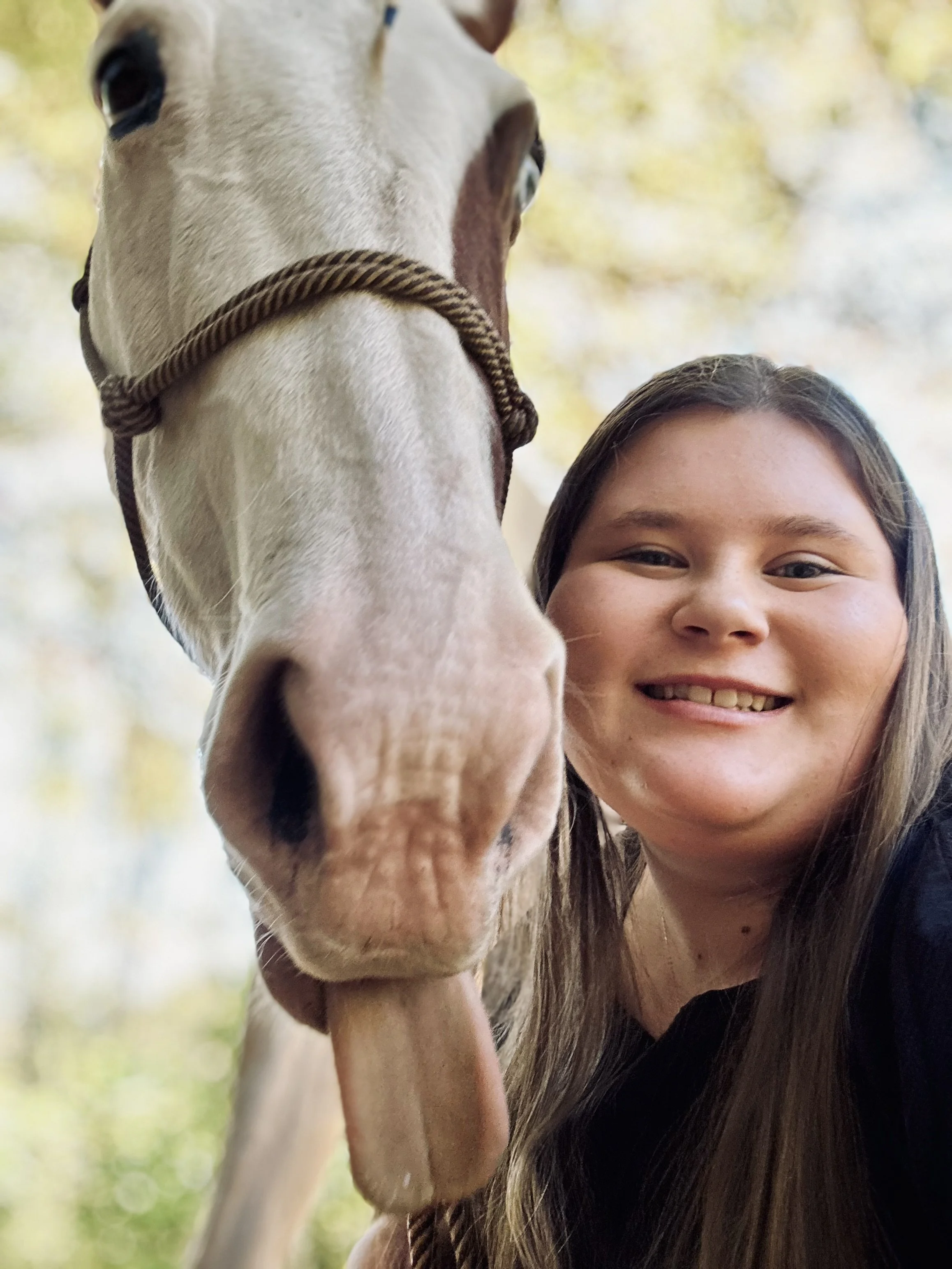 A young woman taking a selfie with a white horse with brown markings during daytime, with blurred trees in the background.